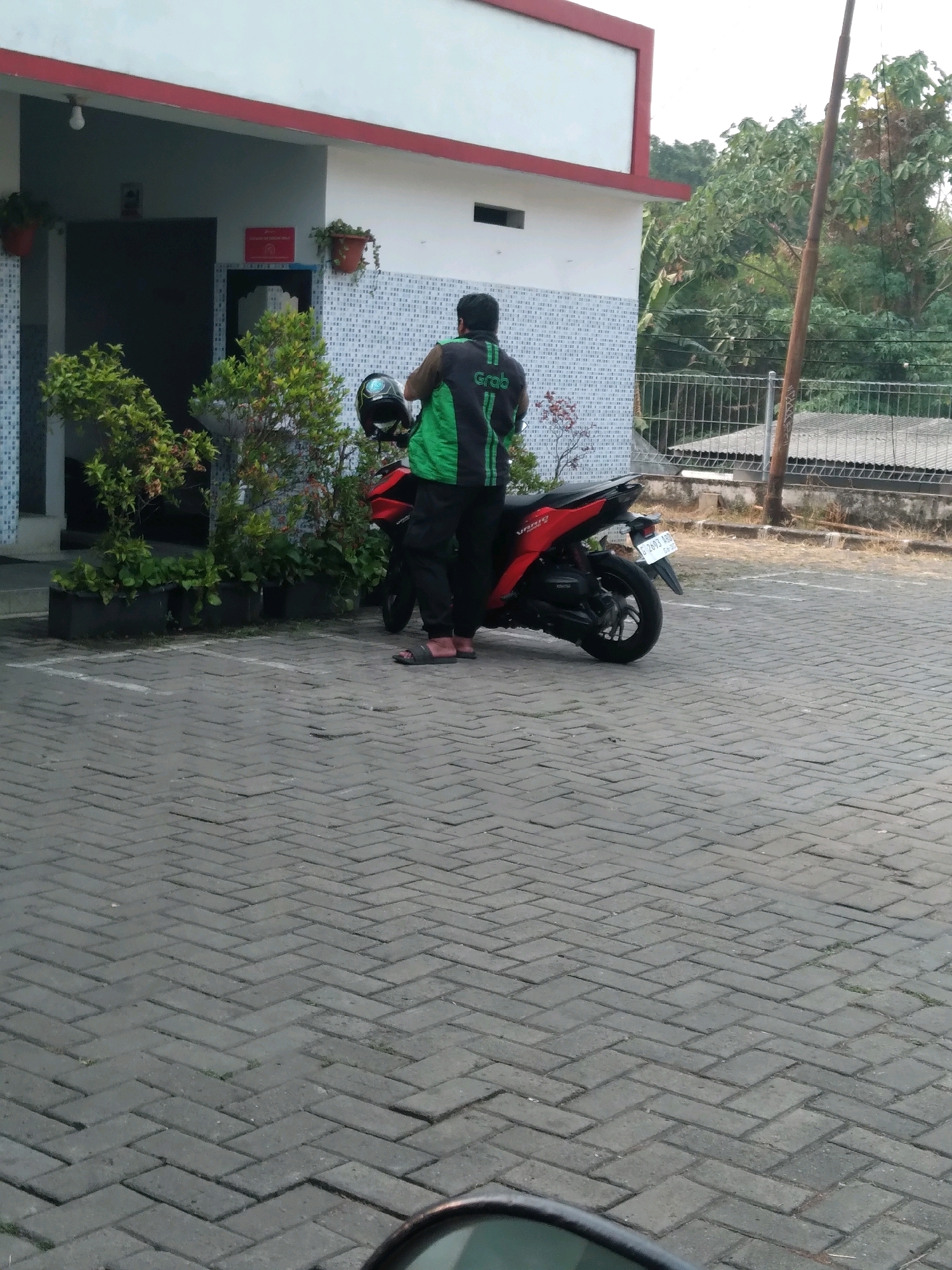 A man wearing a black and green Grab jacket and black pants stands next to a red motorcycle parked on a brick-paved area. The man is facing away from the camera. The motorcycle has a white license plate that reads "B 2603 ABS". In the background, a white building with a red trim and a tiled facade is visible. There are potted plants in front of the building and a metal fence behind the motorcycle. The environment appears to be an outdoor parking area, possibly near a commercial establishment or residential building. The scene is consistent with locations found in the city of South Tangerang, Indonesia, where Grab is a popular ride-hailing service.