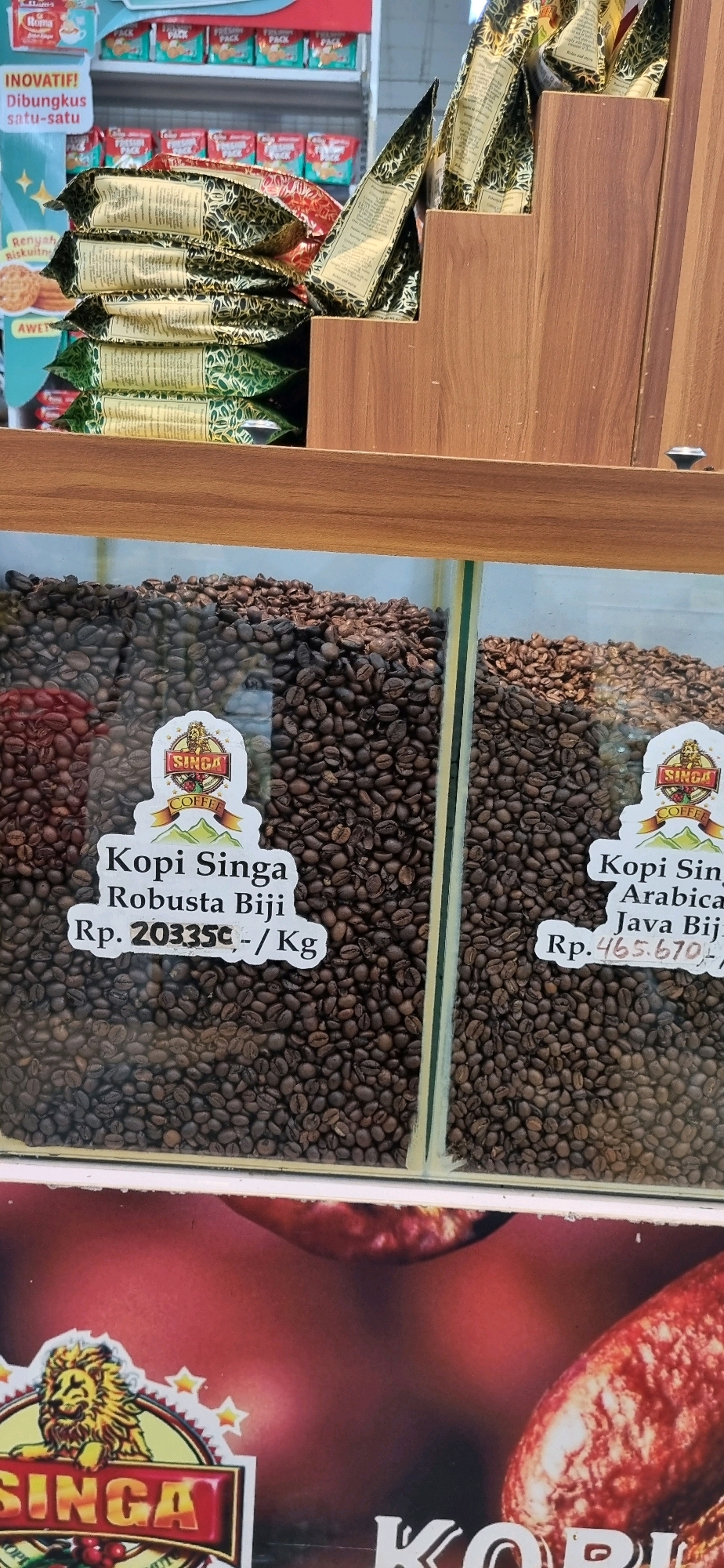 The image shows a display of coffee beans in glass containers at a retail establishment. The coffee beans are dark brown and appear to be roasted. There are two types of coffee beans visible, likely different roasts or varieties, separated by a vertical divider. Each section has a label indicating "Singa Coffee" with sub-labels detailing "Kopi Singa Robusta Biji Rp. 203350,-/Kg" and "Kopi Singa Arabica Java Biji Rp. 465.670,-/Kg". Above the coffee bean display, several packages of what appear to be coffee or similar products are stacked. The overall scene suggests a coffee shop or a section of a larger store selling coffee products. The image quality is moderate, with good lighting.