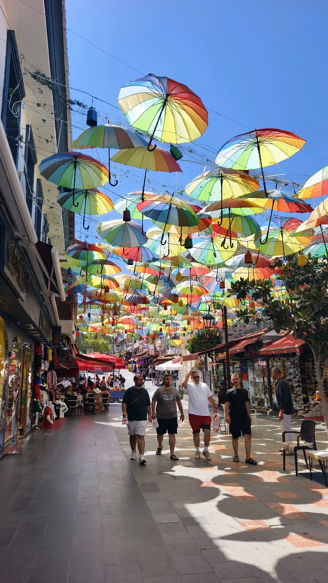 In Kuşadası, Turkey, a pedestrian street is adorned with a vibrant canopy of colorful umbrellas suspended overhead against a clear blue sky. The street is lined with shops on either side, displaying merchandise and awnings, with some outdoor seating areas for cafes. Several people are walking along the paved pathway. In the foreground, four men are walking towards the camera, dressed in casual summer attire. One man in a dark green t-shirt and white shorts, another in a grey t-shirt and black shorts, a third in a white t-shirt and red shorts, and the fourth in a black t-shirt and black shorts. Further down the street, more pedestrians and shoppers are visible, as well as tables and chairs from a cafe. The overall scene depicts a lively and colorful streetscape in a tourist-oriented location.