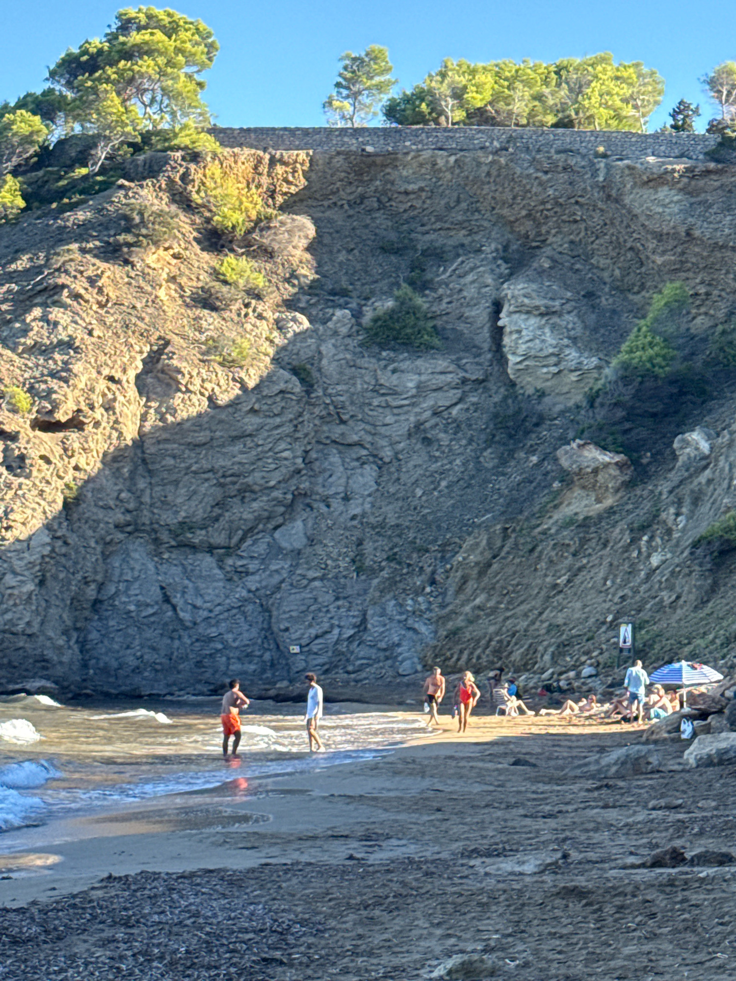 The image depicts a beach scene on a sunny day, likely in Santa Eulària des Riu, Spain, characterized by a rugged, rocky cliff face bordering the sandy shore. Several individuals are present on the beach. Two men are standing in the shallow water, one wearing red swim trunks and the other white shorts. Further up the beach, a group of people are visible, some lying on loungers under a blue and white striped umbrella. Others are standing and interacting. The cliff face, covered in sparse vegetation, rises steeply behind the beach, with a stone wall and trees at its summit. The sea gently laps the shore, and the sand is a mixture of fine grains and small stones. The overall atmosphere is one of relaxation and outdoor activity.