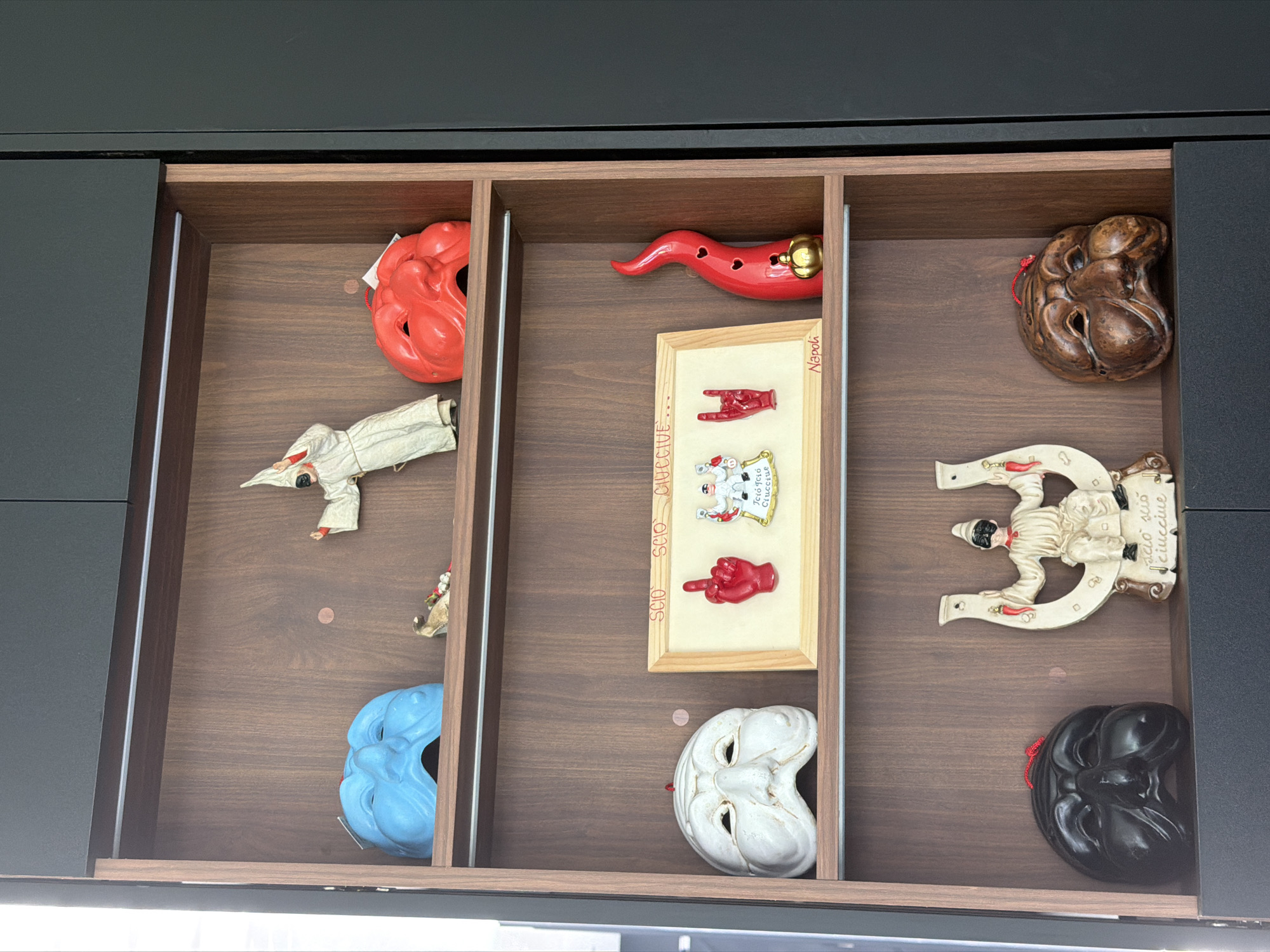 This media file captures an open drawer, neatly divided into three vertical compartments by wooden inserts, showcasing a collection of decorative and cultural items. The drawer and its dividers are made of dark-stained wood.
In the left compartment, a vibrant red, sculpted mask-like object is at the top. Below it lies a small, off-white figure dressed in a pointed hood and cloak, resembling the traditional Italian character Pulcinella. A tiny brown animal figurine is next to it. At the bottom of this section is a bright blue mask with stylized features.
The central compartment features a long, red ceramic chili pepper or horn charm, often known as a "cornicello," at the top. Below it is a small, framed artwork on a light background, displaying two red hand gestures: one forming a "rock on" or "corna" sign, and the other a "fig sign" (mano fica). Text is visible on the wooden frame: "SCIO SCIO CIUCCIUE'" runs vertically on the left side, and "Napoli" is written vertically on the right. Directly below this framed piece is a white theatrical mask with a distinct, expressive face.
The rightmost compartment holds a dark brown, intricately textured, mask-like object at the top. Below it, a larger, off-white horseshoe-shaped decorative piece features another figure resembling Pulcinella, with the phrase "SCIO SCIO CIUCCIUE'" inscribed along its edge. At the bottom of this section, a black theatrical mask with similar features to the blue and white masks completes the display.
The items, particularly the Pulcinella figures, cornicello, and the Neapolitan phrases, strongly suggest a collection of souvenirs or talismans from Naples, Italy. The objects are static, arranged for display or storage. Although the context provided is Puebla, Mexico, the artifacts themselves point to Italian heritage. The lighting is ambient, indicative of an indoor setting with no specific time of day.
