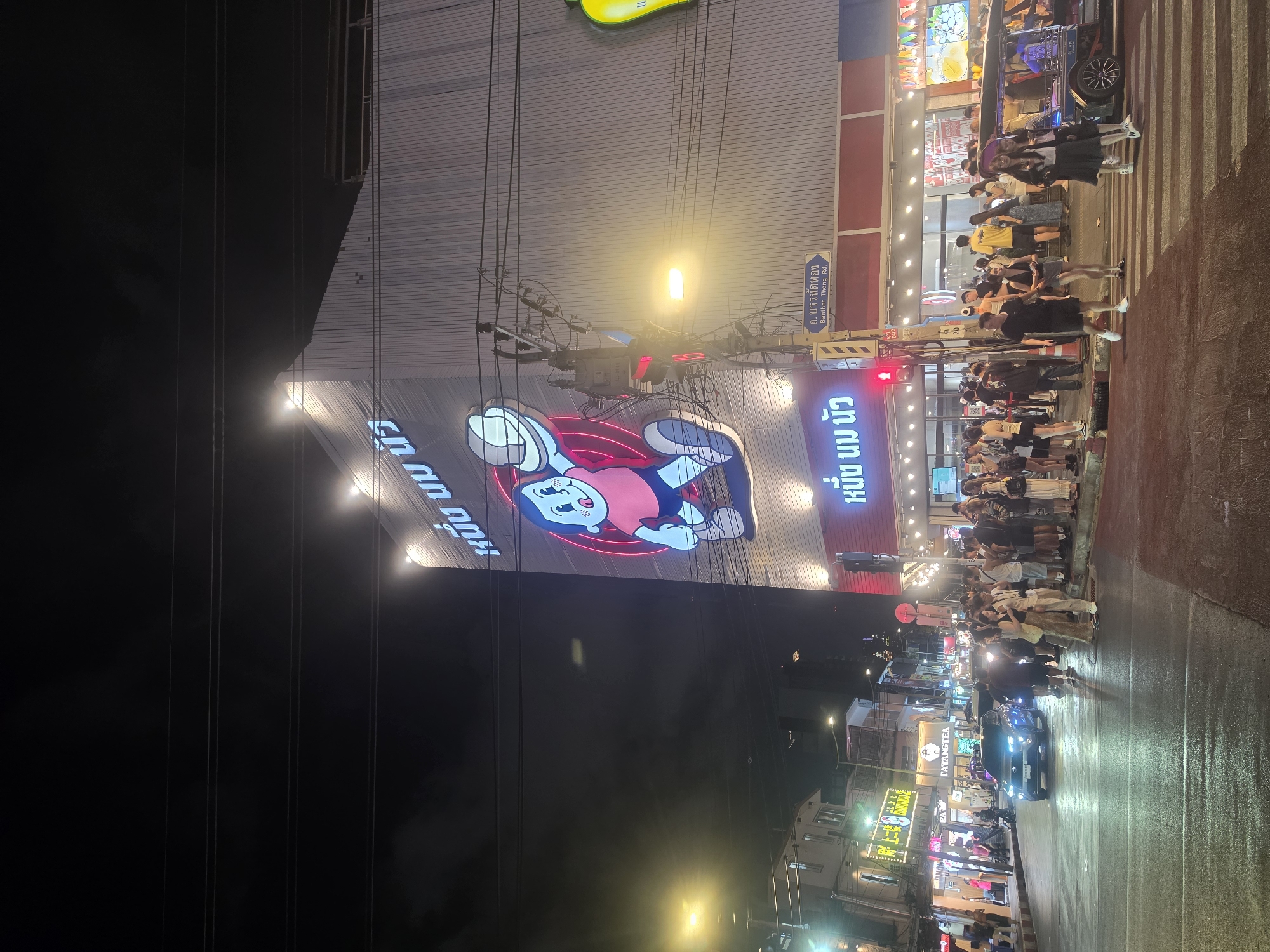 The image captures a nighttime street scene in Bangkok, Thailand, showing a bustling area with shops and pedestrians. The scene is dominated by a large building facade adorned with a sizable, illuminated cartoon character, with text in Thai script alongside it. People are gathered along the sidewalk, many standing on a zebra crossing, indicating a busy pedestrian area. A dark-colored car moves through the street, its headlights illuminating the wet road surface. Various street lights and storefront signs contribute to the brightly lit urban environment. Power lines stretch across the sky, adding to the complexity of the cityscape. The street appears crowded with a mix of locals and tourists enjoying the nightlife. A sign indicates that the location is near Banthat Thong Road.