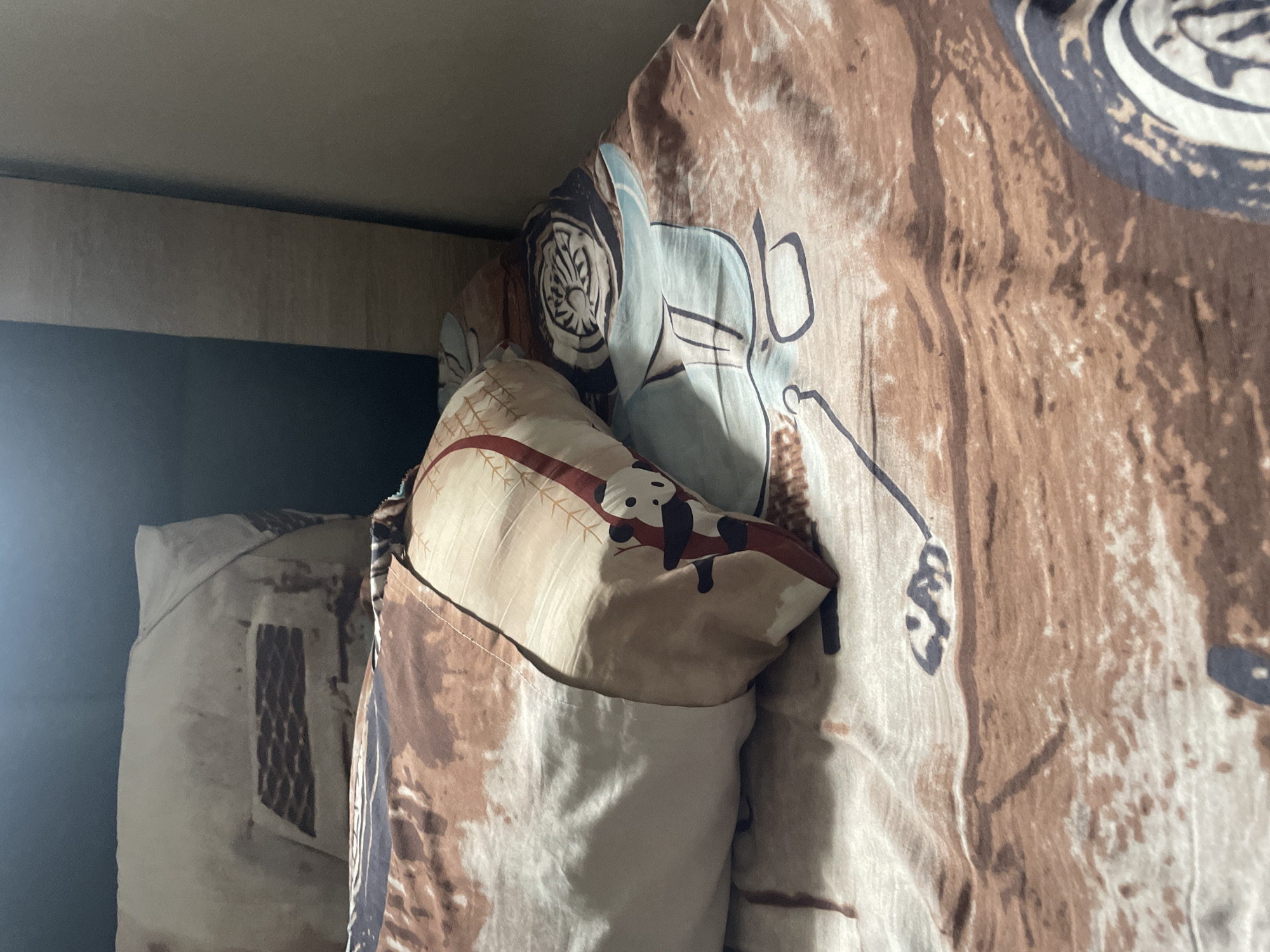 A close-up, angled shot shows a bed with brown and beige patterned sheets. A white pillow, partially covered by a patterned pillowcase, rests on the bed. The pillowcase features a design of a panda climbing a tree. To the right, a portion of a duvet cover displays a light blue graphic, possibly depicting a scene related to golf, with a club and ball. The background is dark, with a vertical wooden panel on the right side of the frame. The setting is a bedroom.