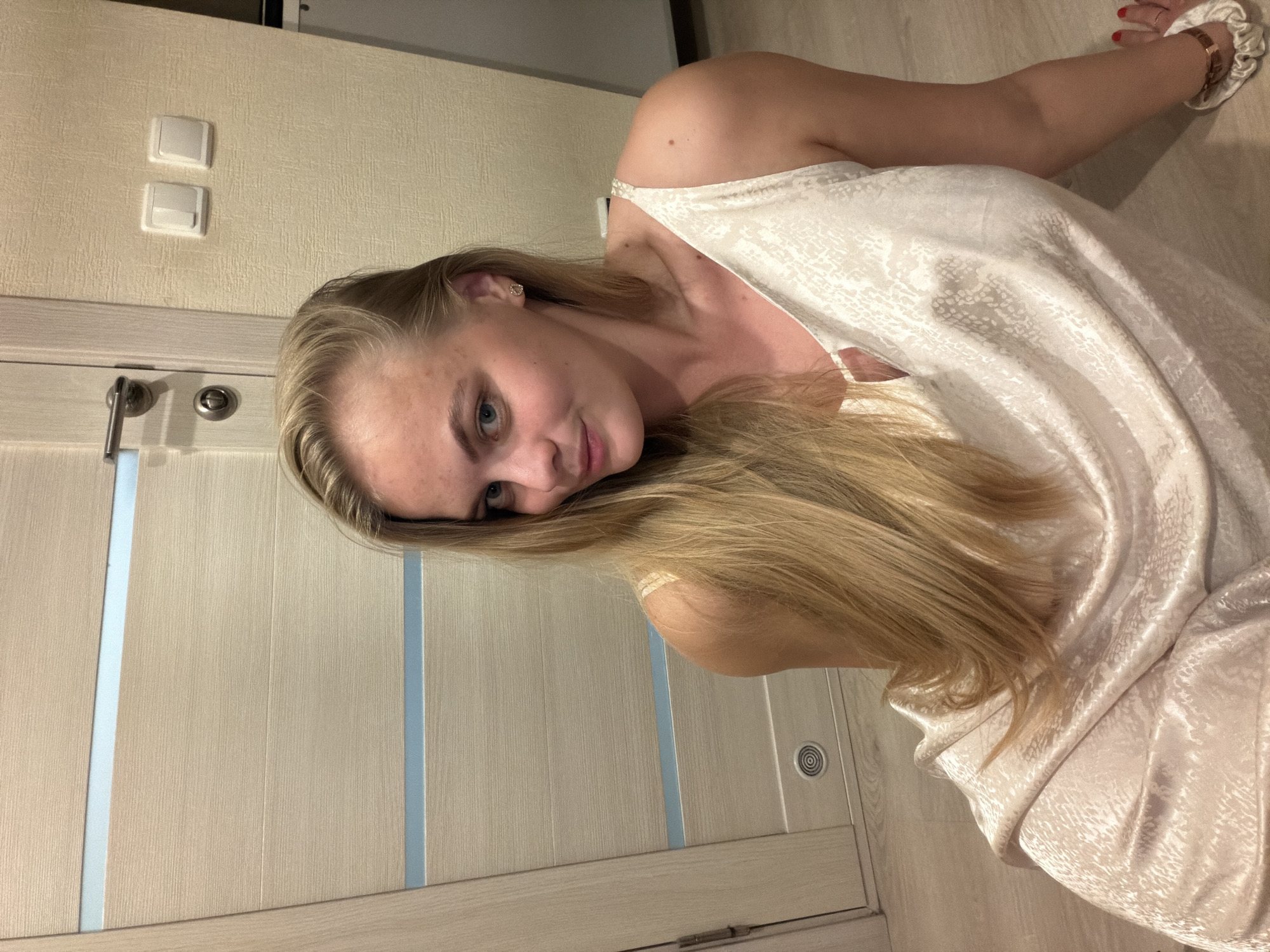 A young woman with blonde hair is pictured indoors. She is wearing a light-colored, patterned slip dress. She is turned towards the camera with a slight smile. Behind her is a light-colored wall with electrical outlets and a door with light blue accents. The location is indoors and there is no indication that it is in Izhevsk, Russia.