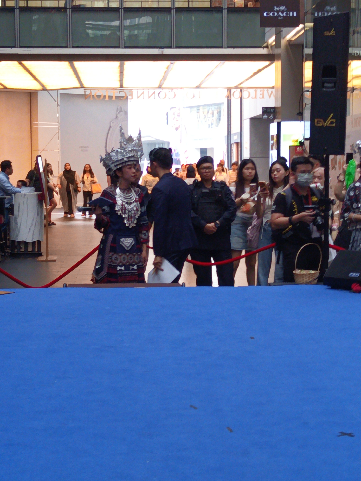 The image shows an indoor event, likely a shopping mall, with a blue carpeted area in the foreground. A young woman wearing a traditional ethnic costume with a large, ornate headdress and silver jewelry stands in the middle of the frame. She is holding a book or tablet and appears to be speaking or presenting. Behind her, several people are gathered, including individuals in security uniforms, shoppers, and possibly event staff. In the background, there are store signs, including one for "COACH" and another with a mirrored "CONNECTION" text. The setting suggests a cultural or promotional event taking place in a public space, possibly within a commercial district in Kuala Lumpur, Malaysia, given the "COACH" signage which is prevalent in many upscale malls in the city. The overall scene captures a moment of public engagement or performance.