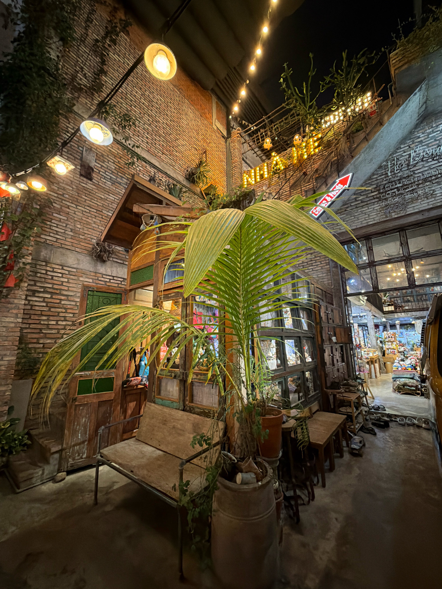 The image depicts an outdoor seating area in front of a building at night in Nha Trang, Vietnam. The scene features a rustic aesthetic with exposed brick walls, wooden doors and windows, and a variety of plants. A long, wooden bench with metal legs is positioned centrally, facing the building. Several smaller wooden tables and chairs are arranged to the right of the bench, with some items that appear to be shoes scattered on the ground.
The building's facade is characterized by brickwork and large, multi-paned windows. An entranceway with a wooden door and a small gabled roof is visible to the left of the bench. Above the entrance, a large palm frond extends into the foreground, partially obscuring the view. String lights are visible hanging from the ceiling and along the upper portion of the building, illuminating the area.
In the background, a sign with red and white lettering, including an arrow pointing to the right, is mounted on the wall. Another illuminated sign with yellow lettering spelling out "RIFF..." is also visible higher up on a balcony or upper level. The interior of the building is dimly lit, with displays of merchandise visible through the windows, suggesting a shop or restaurant. The ground is paved with concrete. The overall impression is one of a bohemian or eclectic urban setting.
