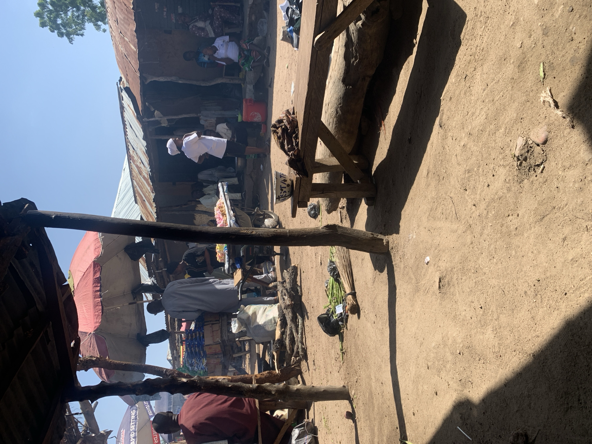 The image depicts a bustling market scene in Boi A, Nigeria. The scene is outdoors, with a dirt ground and simple, rustic structures that suggest a traditional marketplace. The overhead lighting and long shadows indicate it is daytime, likely late morning or early afternoon. The weather appears to be clear and sunny.
Several individuals are visible, dressed in everyday clothing, some wearing white garments and head coverings, suggesting participation in a religious or cultural observance. Some people appear to be vendors, while others are customers or passersby. The activity suggests a typical market day, with goods being displayed and transactions likely taking place.
Objects in the scene include various stalls with goods, a large red container, a broom made of natural materials, and wooden structures that serve as part of the market's infrastructure. One stall is covered by a large, red umbrella. A large tree trunk has been incorporated into a wooden bench or table.
Notable details include the corrugated metal roofing of some structures, indicative of makeshift or semi-permanent market stalls. The overall atmosphere seems lively and busy, typical of a community gathering point. There are no overt signs of distress or negative emotions; the scene appears to be one of everyday commerce and social interaction.
There is some visible text, though it is difficult to read clearly. One sign, partially obscured, appears to have English words such as "SETTING" and "RAPID," which could relate to business names or product advertisements.