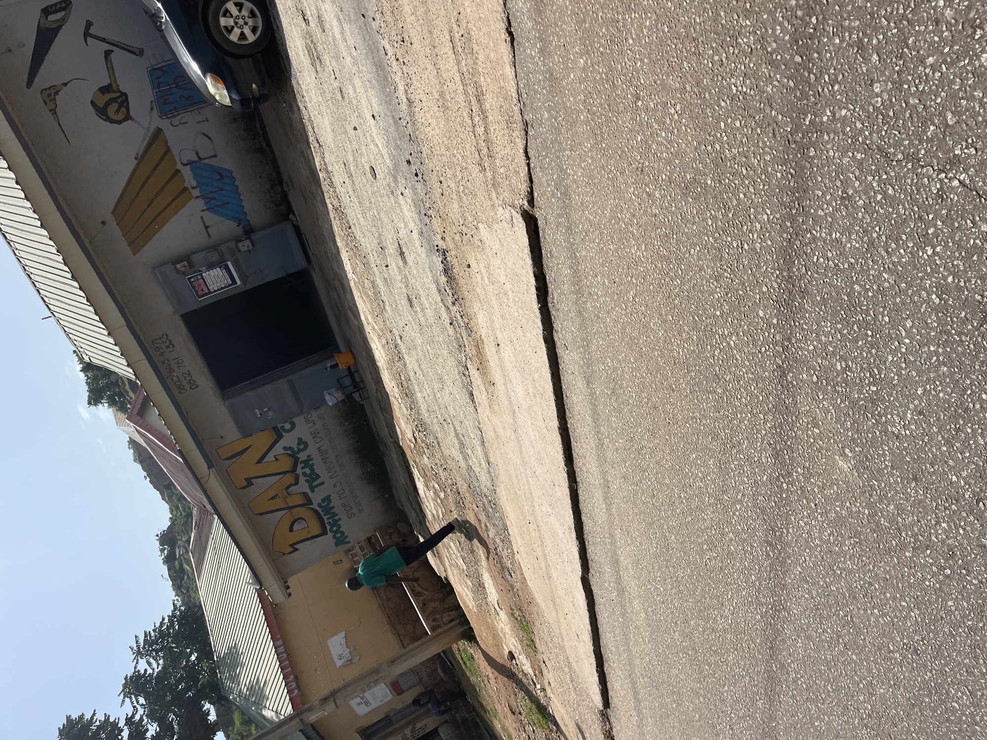 The image depicts a street scene in Gura Top, Nigeria. A man in a green shirt and dark pants is walking away from a building. The building, identified as "DAN ROOFING TECH. &... CAR WASH", has a mural on its wall featuring various tools and what appears to be corrugated metal sheets. A black sedan is parked in front of the building. The street in the foreground shows signs of wear and tear, with cracks and uneven surfaces. The background reveals a hilly landscape with trees and more buildings, suggesting a semi-urban or rural environment. The sky is clear and blue, indicating a bright, sunny day.