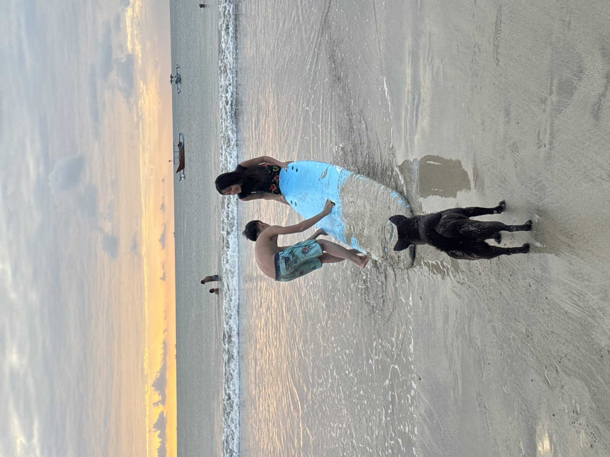 In Kuta Selatan, Indonesia, two young individuals, a boy and a girl, are engaged with a surfboard on a beach at dusk. The boy, wearing patterned swim trunks, is pushing the surfboard forward into the shallow water. The girl, dressed in a dark floral-patterned top and a floral skirt, is holding the front of the surfboard. A black dog stands on the wet sand between the viewer and the surfboard, facing away from the camera and towards the ocean. In the background, gentle waves roll onto the shore, and further out, several boats are visible on the calm sea. The sky above is filled with soft clouds tinged with the warm hues of the setting sun. The sand is wet and reflective, showing ripples from the receding tide.