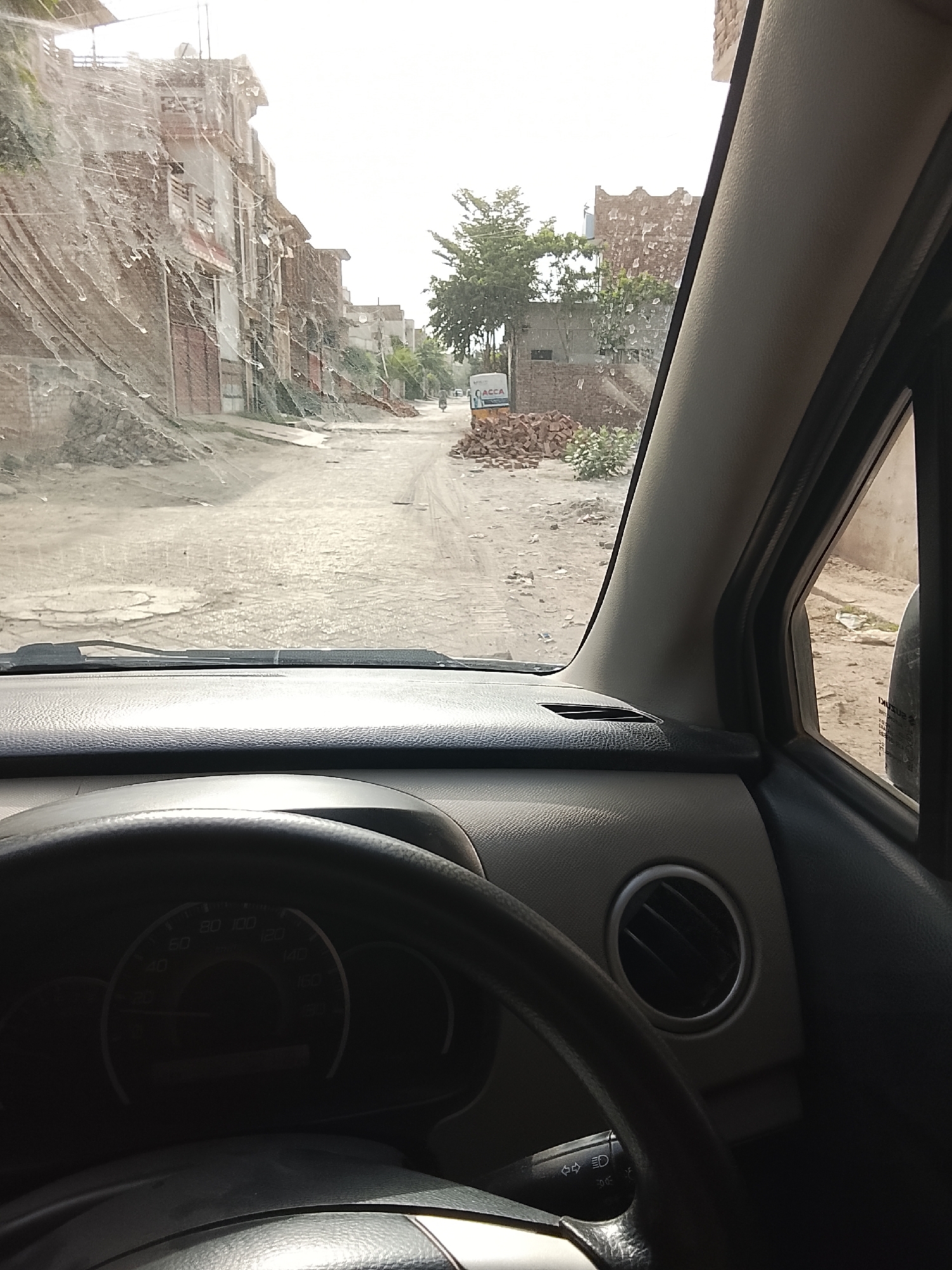 The image shows a view from inside a car, looking out onto a street. The street is unpaved and appears to be in a state of disrepair, with piles of bricks or rubble scattered along the roadside. Buildings line both sides of the street, appearing old and made of brick. A tree can be seen in the distance. A motorcycle is traveling down the street. The car's dashboard and steering wheel are visible in the foreground. The location is Gulgasht Colony, Pakistan.