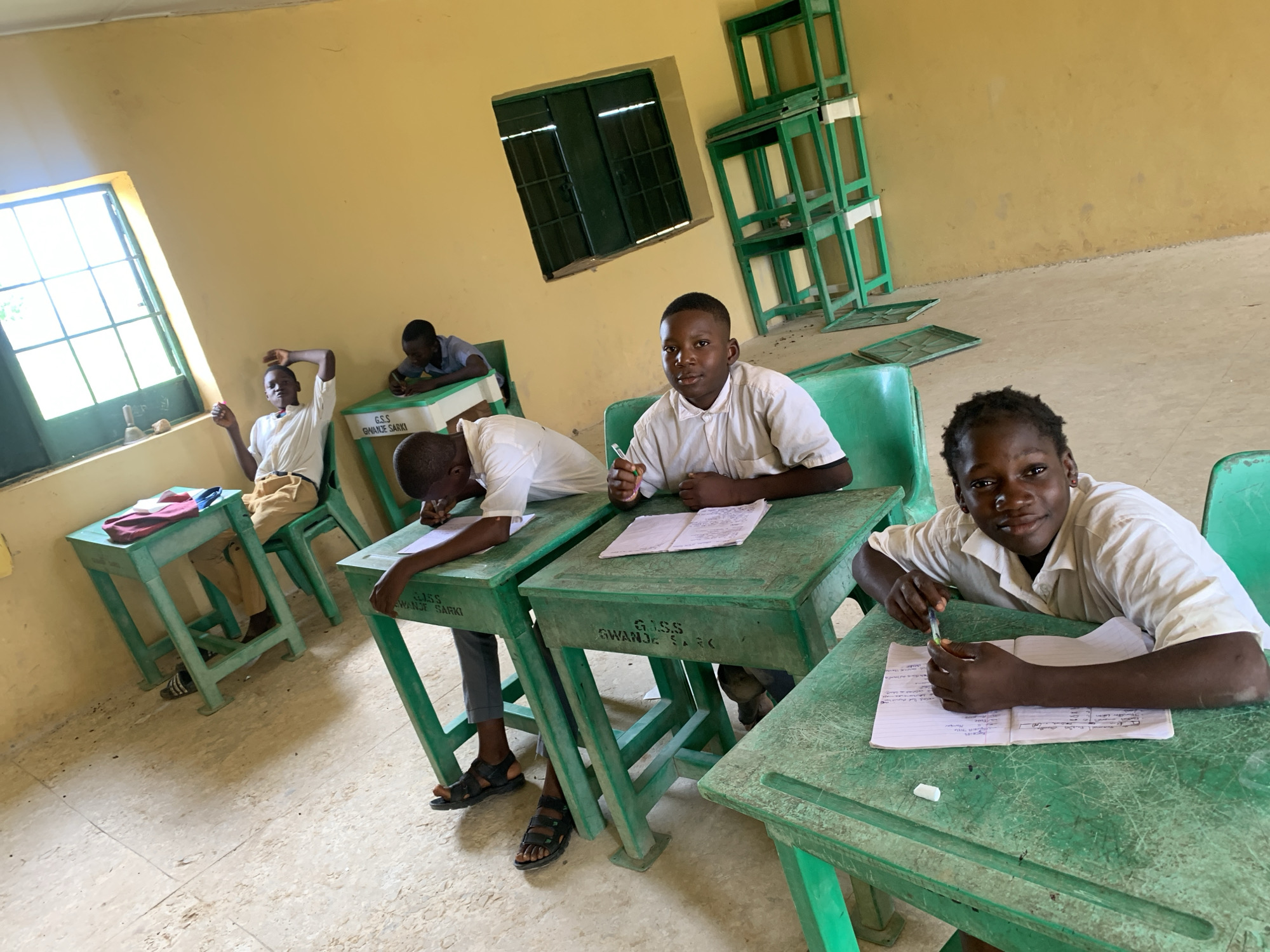 The image depicts an indoor scene, likely a classroom, in Akwanga, Nigeria. Four young individuals, presumably students, are seated at green desks. The room has yellow walls and a concrete floor. Sunlight streams in from a large window with green-framed panes. In the background, stacks of identical green desks and chairs are piled against the wall.
The student in the foreground, on the right, is looking directly at the camera, smiling slightly, and holding a pen. They are wearing a white collared shirt. In front of them is an open notebook with handwritten notes and diagrams. A white eraser is also visible on the desk.
To the left of the foreground student, another student is leaning forward, writing in an open notebook. This student is wearing a white shirt and dark pants. Their desk is also green and has "G.S.S. GWANJE SARKI" inscribed on it.
Further to the left, a third student is seated at a desk, with their head bowed as if concentrating on writing or reading. They are wearing a white shirt and tan pants.
In the background, near the window, a fourth student is seated at a desk, leaning back with their right arm raised and bent at the elbow, as if stretching or resting. This student is wearing a white shirt and tan pants.
The overall impression is of a functional, albeit somewhat basic, educational environment.
