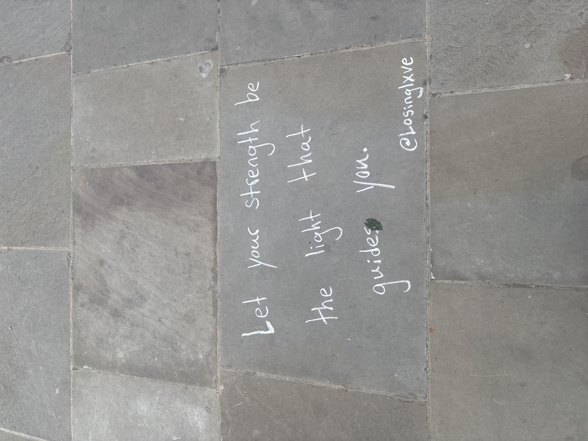 The image displays a close-up, ground-level view of grey paving stones, typical of a sidewalk or public area in London. The pavement consists of multiple irregularly shaped and sized flagstones, varying slightly in shade and texture.
Written prominently in white chalk across several adjacent slabs is an uplifting message. The text, oriented vertically within the frame, reads: "Let your strength be the light that guides you." Below this inspirational quote, a social media handle, "@losinglxve," is also inscribed in chalk. A small green leaf is visible resting on the pavement near the word "guides."
The lighting suggests it is daytime, and the chalk is clear and intact, indicating dry weather conditions. There are no people or other distinct objects present, keeping the focus entirely on the chalk message against the backdrop of the urban hardscape.