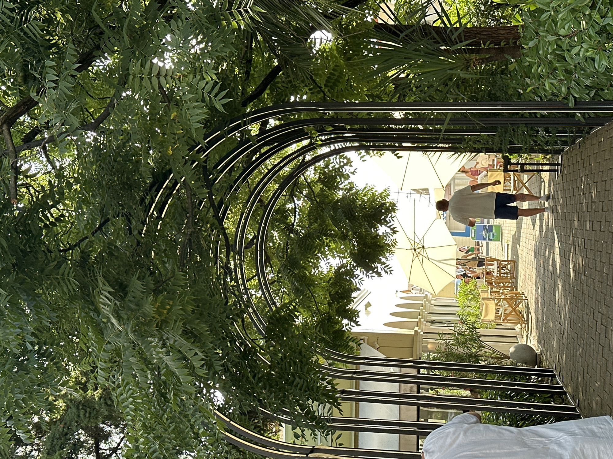 The image depicts an outdoor seating area with a man walking away from the camera down a brick pathway. He is wearing a white t-shirt and blue shorts. The pathway is lined with green foliage and a metal archway structure covered in leaves. In the background, there are tables and chairs set up outdoors, some with umbrellas for shade. Several people are visible in the background, seated at tables. The architecture in the background appears to be a building with arches. The setting suggests a cafe or restaurant in a park-like environment. The image is taken on a sunny day, with sunlight filtering through the leaves. This scene is located at the Arboretum Botanical Garden in Sochi, Russia, a popular tourist attraction.