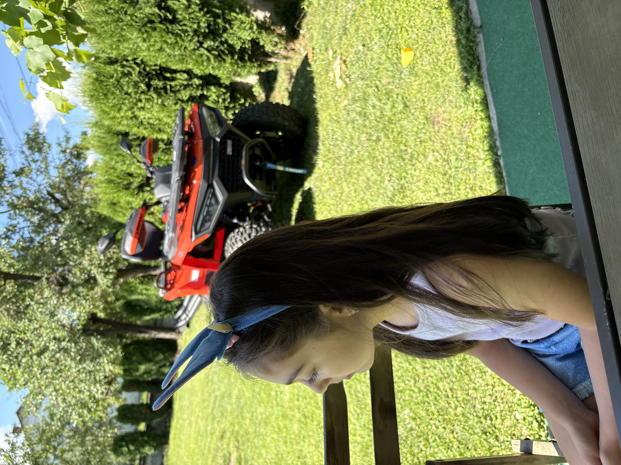 A young girl with long, dark hair is looking down, her face partially obscured. She is wearing blue denim shorts and a light-colored tank top, with a blue headband adorned with fluffy bunny ears on her head. The girl appears to be sitting on a wooden surface, possibly a bench or porch, with a dark railing in the foreground.
Behind her, the scene unfolds in a sunlit backyard. A vibrant orange ATV is parked on the green grass, its tires resting on the lawn. The ATV has black accents and what appears to be a roll cage. Lush green trees and bushes form a natural backdrop, with some leafy branches visible in the upper left corner. A clear blue sky with a few wispy clouds suggests it is daytime, likely a sunny day. The overall setting is a residential backyard, possibly in a rural or suburban area given the abundance of greenery and the presence of the ATV, common for outdoor recreation. The atmosphere seems relaxed and casual, indicative of a warm day. There is no visible text in the image.