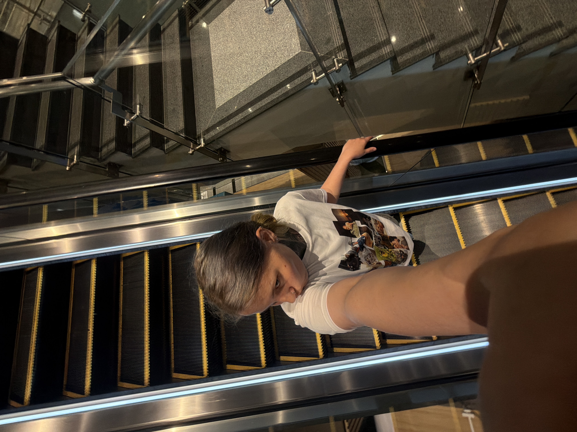 A young person is captured in a selfie-style shot while riding an escalator inside a modern building in Đà Nẵng, Vietnam. The individual, with brown hair partially tied back, is wearing a white t-shirt featuring numerous small printed images. A nose piercing is visible on their face, which holds a neutral expression as they look directly at the camera. Their extended arm, holding the camera, is prominent in the lower right foreground.
The escalator features distinct black steps outlined with yellow accents, accompanied by a polished metal handrail. The perspective suggests a descent. In the background, a dark, multi-tiered staircase is visible, ascending alongside the escalator. This staircase area is delineated by contemporary glass railings supported by metallic fixtures and posts. Beyond the glass, textured grey wall or flooring surfaces are apparent. The lighting is generally subdued, contributing to an indoor, contemporary atmosphere. The architectural elements, including the glass, metal, escalator, and staircase, suggest a modern public or commercial space, such as a shopping mall, hotel, or convention center. No legible text is visible on the person's attire or within the environment.