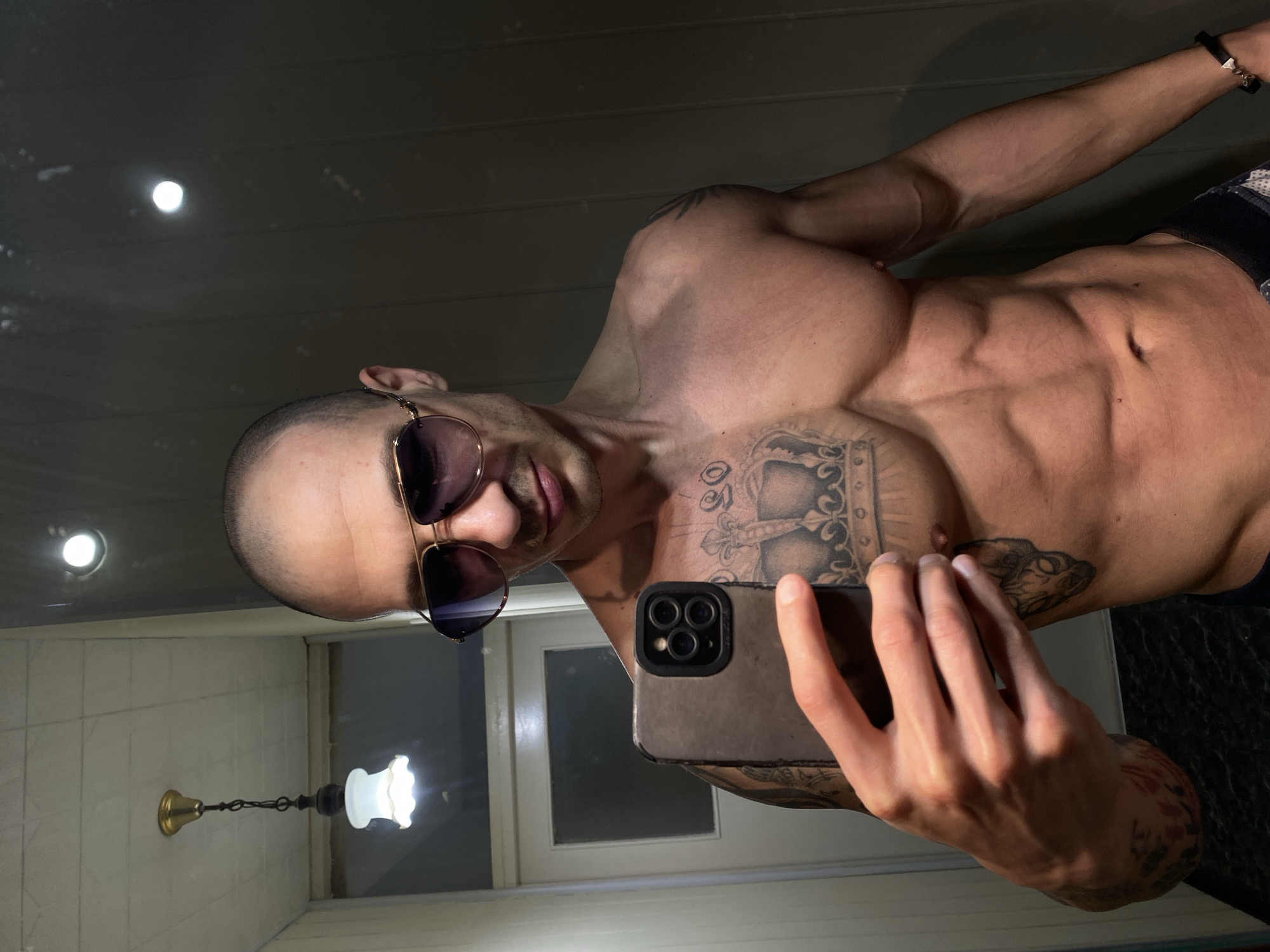 A shirtless man with a shaved head and sunglasses is taking a selfie in a room. He has several tattoos on his chest and arms. He is holding a cell phone in his right hand. In the background, there is a light fixture hanging from the ceiling. The exact location in the city of Pleven, Bulgaria cannot be determined from the image alone.