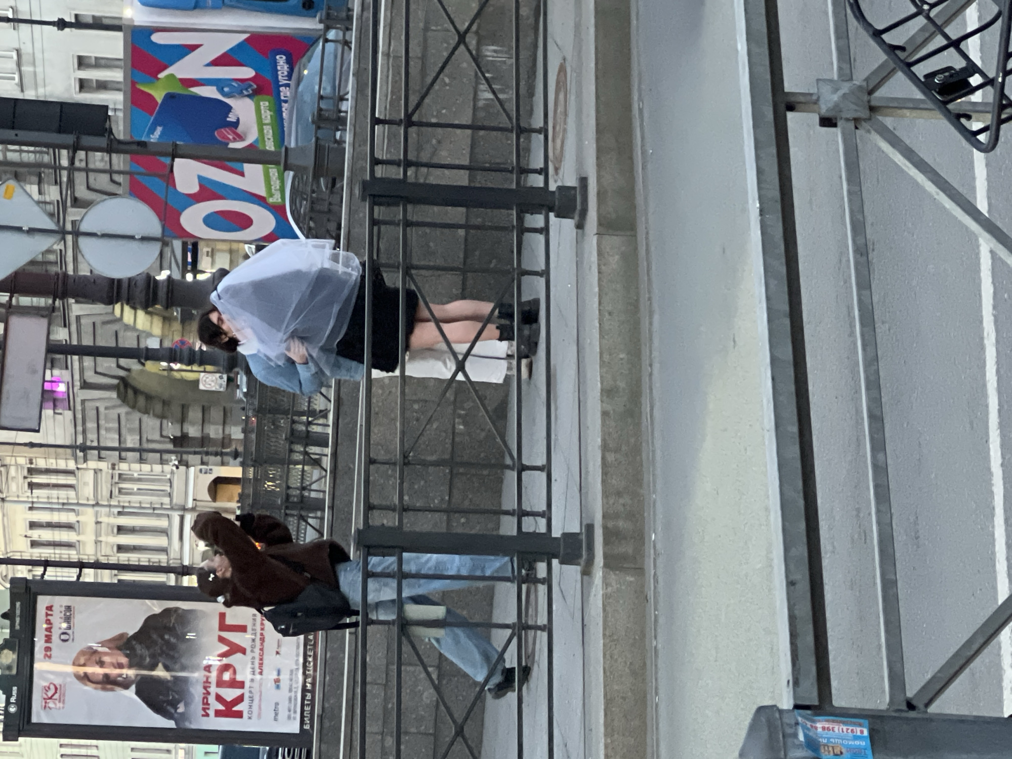 Here is a description of the image:
The photograph shows a street scene in Saint Petersburg, Russia. In the foreground, there are two people standing behind a metal fence. One person is wearing a brown jacket and jeans, and is taking a photo with their phone. The other person is wearing a light blue shirt, a white translucent covering, and a black skirt. They are leaning against the fence. Behind the fence is a sidewalk and a street with car traffic. Buildings are visible in the background. Also a billboard poster of Russian singer Irina Krug on a street lamp post with the concert date March 29. An ad for OZON is also shown above.