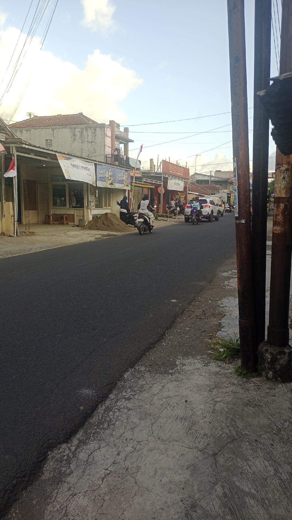 The image depicts a street scene in Pakemitan, Indonesia, likely during the daytime. The road is paved with asphalt, and a concrete sidewalk runs alongside it, showing signs of cracking and wear. Several buildings line the street, appearing to be shops or small businesses, with some featuring awnings and signage. One sign reads "SWEET PANDA."
In the foreground, the road is empty except for a pile of sand near the sidewalk. Further down the street, several people are present. Two individuals are on a motorcycle, with one driving and the other as a passenger. A white SUV is also visible, as is another motorcycle. People are seen walking or standing near the businesses.
The sky above is partly cloudy, with a mix of blue sky and white clouds. Several electrical wires are strung across the scene, suspended by poles. Two large, dark, vertical poles are prominent on the right side of the image, appearing to be utility poles. One of these poles has some graffiti or markings on it. The overall environment suggests a commercial or residential street with ongoing activity.
