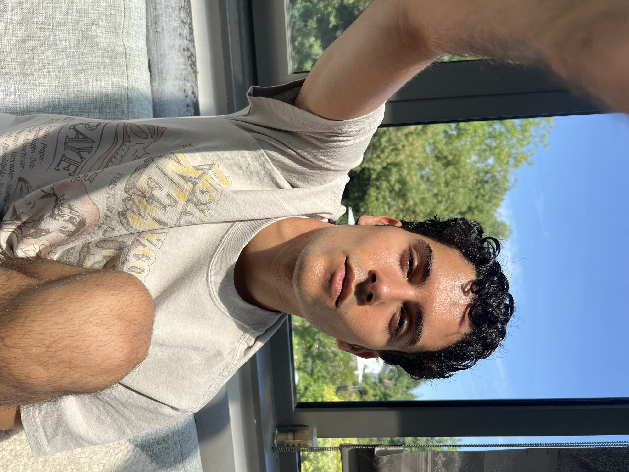 A young man with dark curly hair and olive skin is taking a selfie. He is sitting indoors, with a window behind him that looks out onto trees and a clear blue sky. The man is wearing a light-colored t-shirt with a graphic print. His right arm is extended, holding the camera. He is looking directly at the camera, with a slight smile. The lighting suggests it is daytime and sunny. The background shows a glimpse of a city, with a building visible through the trees.