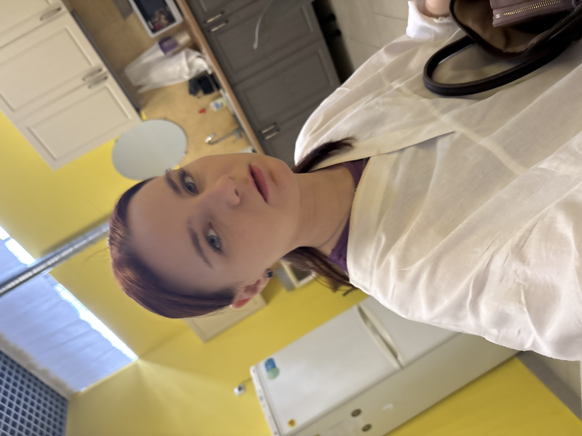 A person with reddish-brown hair and blue eyes is partially visible in the frame. The person is wearing a white garment. A partially visible handbag is also in the frame. The environment includes white and grey cabinets, and yellow walls. A refrigerator is also present. The location appears to be inside a building, possibly a kitchen or clinical space, but it is unclear if it is in Sinyavino, Russia.