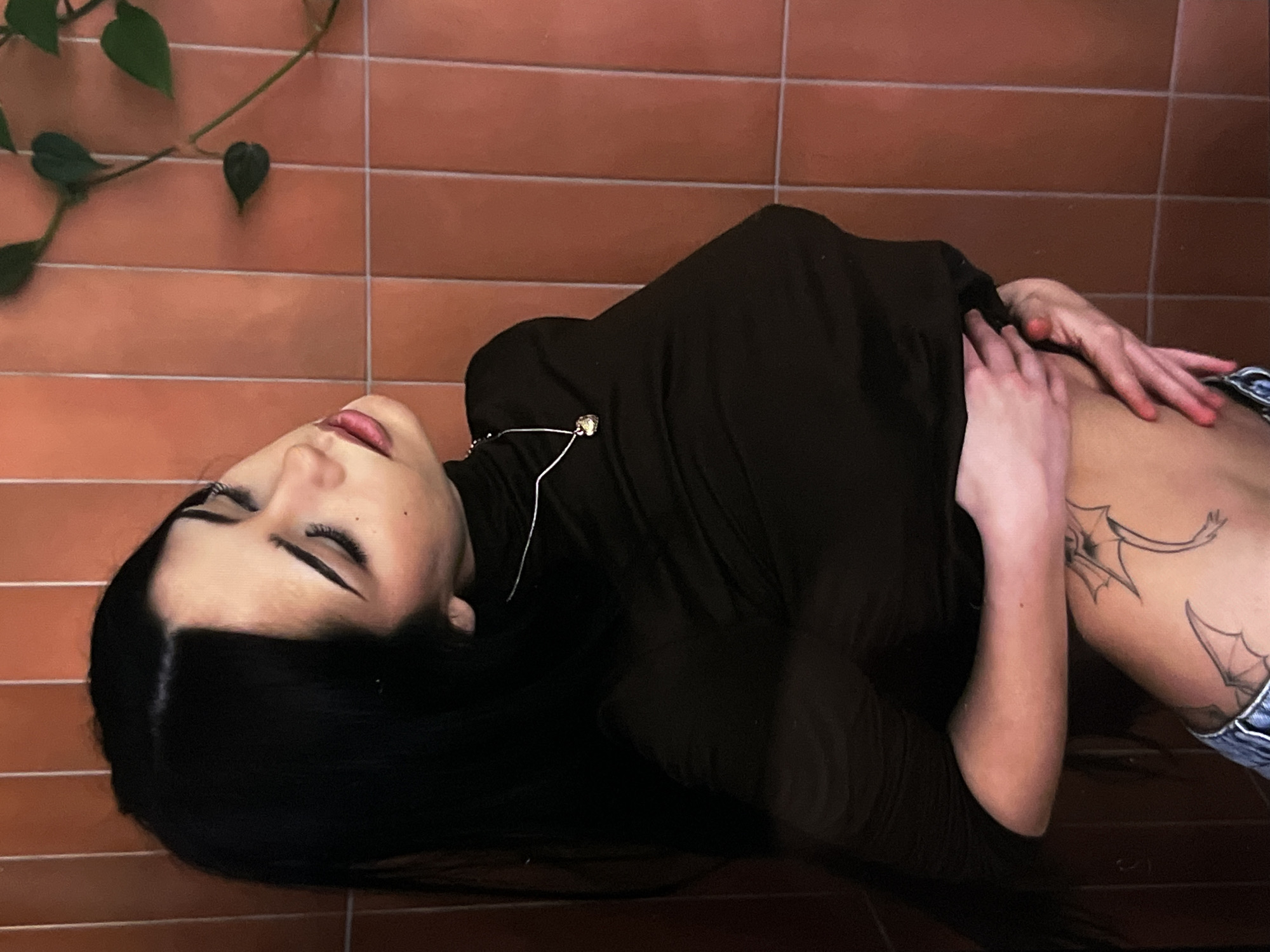 A young woman with long black hair, wearing a dark turtleneck and jeans, is pictured in a studio-like setting with terracotta-colored tiled walls. She has a tattoo on her hip. Her head is tilted back, and her eyes are closed, with makeup visible on her eyelids and lips. A delicate necklace with a heart-shaped pendant adorns her neck. Her left hand is placed on her abdomen, lifting her shirt slightly to reveal the tattoo. A vine with green leaves hangs on the wall behind her. The lighting is dim, casting shadows on her face and body.
