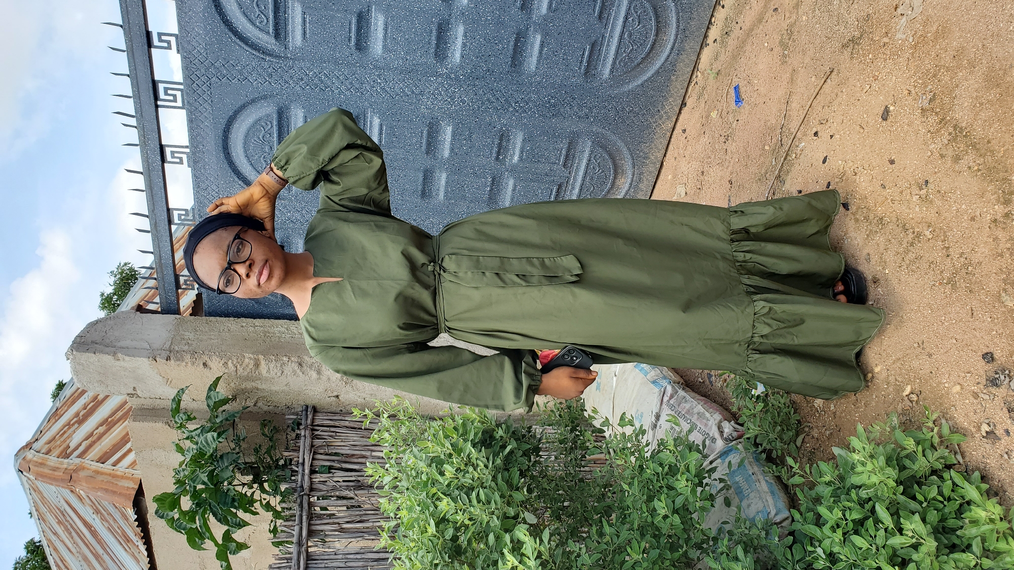A young woman wearing glasses, a black headscarf, and a long olive-green dress stands in front of a grey metal gate and a textured concrete wall. Her left hand is raised to her head, and her right hand holds a smartphone. To the left of the woman are a wooden fence, a leafy green bush, and two large sacks of cement. Overhead, the sky is overcast. The scene appears to be in an outdoor area in Jalingo, Nigeria.