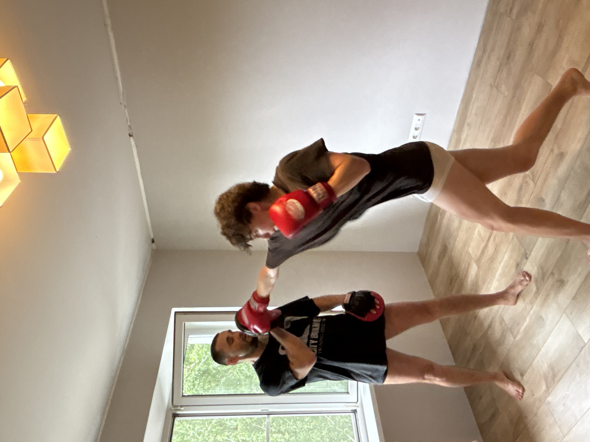 The image depicts two individuals engaged in a boxing training session indoors. One individual, appearing to be an instructor, stands on the left, wearing a black t-shirt and black shorts, with red boxing gloves on his hands. He holds a striking pad, also red, in his left hand, and his right hand is extended forward. The other individual, a younger person with curly hair, stands on the right, wearing a black t-shirt and white briefs, also with red boxing gloves. This person is in the motion of throwing a punch. The environment appears to be a room with light-colored walls, a wooden floor, and a large window with greenery visible outside. A modern, cube-shaped light fixture is visible on the ceiling. The presence of a power outlet on the wall suggests an indoor setting. There is no indication that this scene is located in the city of Chaikovsky, Russia.