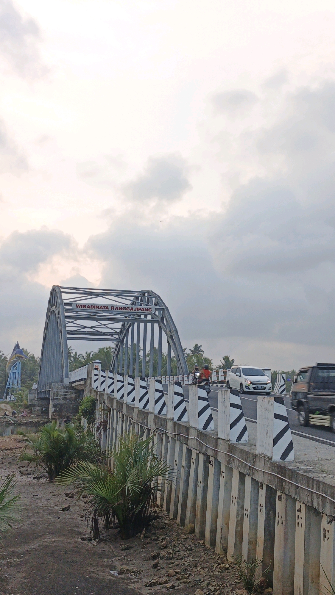 The image captures the "Wiradinata Rangganjipang" bridge in Pangandaran, Indonesia, under an overcast sky. The bridge features a distinctive arched metal structure with the name prominently displayed. On the left side of the bridge, a large blue statue of a marlin is visible. The road on the bridge is actively used, with a white car, a black SUV, and a motorcycle with a rider and passenger in motion. The bridge's railing is adorned with black and white diagonal stripes. The surrounding environment includes palm trees lining the horizon, and the ground beneath the bridge is a mixture of mud, small stones, and sparse vegetation. The overall scene depicts a functional piece of infrastructure in a tropical setting.