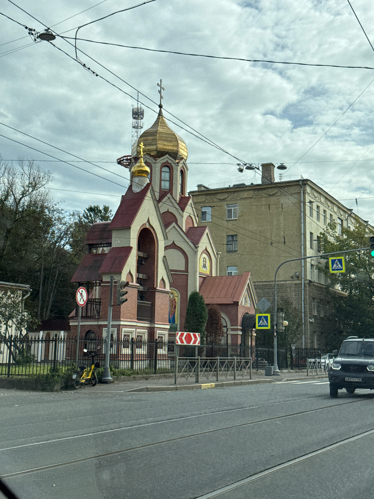 A street scene in Saint Petersburg, Russia, features a small, red-brick Eastern Orthodox church with a golden dome. The church is situated near a multistory apartment building and several trees. Traffic signals and pedestrian crossing signs are visible. A black SUV is driving on the street, and a yellow moped is parked on the sidewalk. Overhead power lines crisscross the sky, and a telecommunications tower is visible in the background. The scene appears to be captured from a vehicle, as suggested by the perspective.