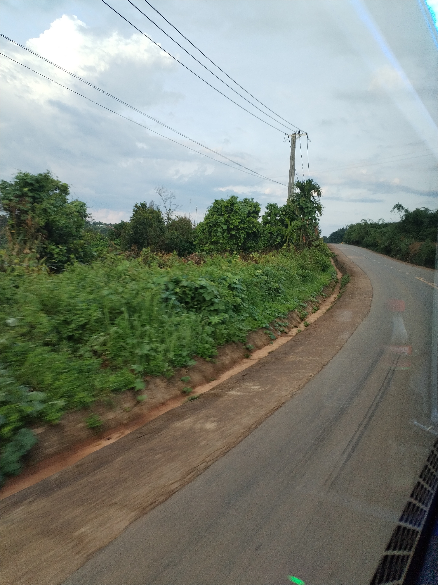 The image depicts a paved road curving through a landscape of dense green vegetation under an overcast sky. A power pole stands along the roadside with multiple power lines extending across the frame. The view appears to be taken from a moving vehicle, indicated by the blurred foreground, an unclear reflection and a vehicle vent. The road is bordered by a dirt shoulder transitioning to lush foliage. The location is Pờ Y, Vietnam.