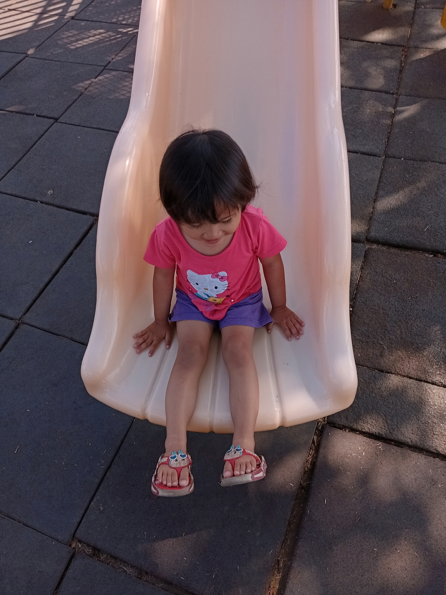 A young child, likely a girl, is sitting on a light-colored plastic slide at a playground. She is wearing a pink t-shirt with a graphic of Hello Kitty and purple shorts. On her feet, she has red sandals with decorative butterfly details. The child is looking down with a slight smile, suggesting she is enjoying herself.
The playground surface appears to be made of interlocking dark gray rubber tiles, which is a common safety feature for playgrounds. Shadows from an overhead structure, possibly a fence or part of a larger playground apparatus, are cast across the tiles, indicating it is daytime. The lighting suggests the sun is at an angle, possibly late afternoon.
The scene is set outdoors, likely in a park or playground area. The specific mention of Purworejo, Indonesia, provides geographical context. There is no visible text in the image. The overall mood is cheerful and relaxed, capturing a simple moment of a child playing.