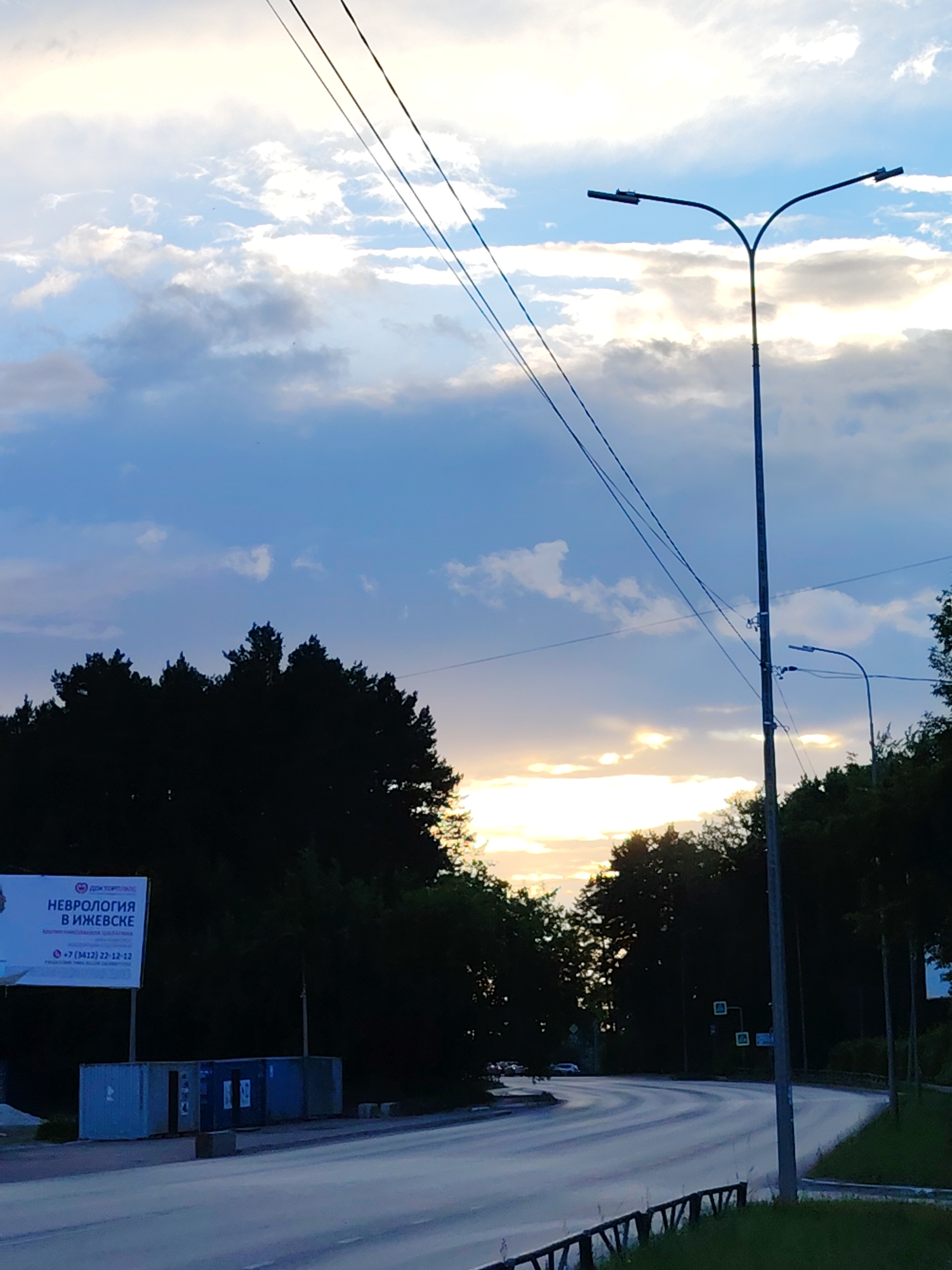 The image depicts a scenic view of a road curving through a wooded area, likely in Chaikovsky, Russia. The time of day appears to be sunset or sunrise, as indicated by the warm glow of the sun breaking through a cloudy sky. The sky is filled with dramatic clouds in shades of blue, white, and gray, with streaks of yellow and orange where the sun's rays are most intense.
Dominating the foreground is a tall lamppost with two lights, its silhouette stark against the sky. Several power lines also crisscross the frame, adding to the urban-meets-nature aesthetic. On the left side of the road, a billboard stands prominently, displaying text in Russian. The visible text reads "ДОКТОР ТОЛСТОЙ" and "НЕВРОЛОГИЯ В ИЖЕВСКЕ," suggesting a medical clinic specializing in neurology in Izhevsk. Below this, a phone number is visible: "+7 (3412) 22-12-12." In front of the billboard, there are several large, rectangular containers, possibly for storage or waste disposal.
The road itself is smooth and appears to be well-maintained, with white lines marking the lanes. The trees bordering the road are dark and dense, forming a thick silhouette against the brighter sky. On the right side of the road, more trees are visible, along with a glimpse of what might be a grassy verge and a low fence. In the distance, a few cars can be seen on the road, suggesting ongoing traffic. The overall mood of the image is peaceful and slightly serene, with the natural beauty of the sunset contrasting with the man-made elements of the road and lampposts.