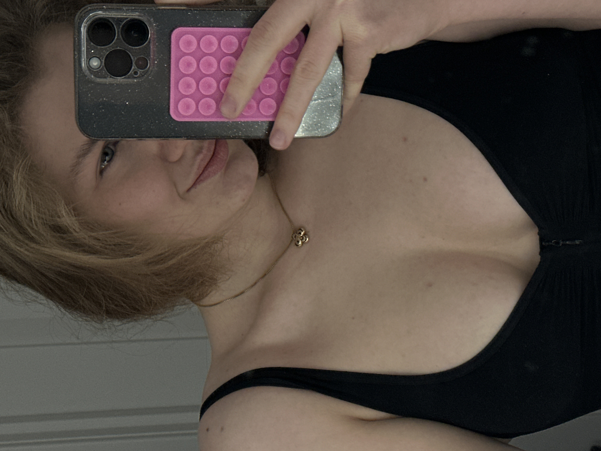 A young person with fair skin and blonde hair is taking a selfie, holding a smartphone with a black glitter case and a pink circular bubble pad. The person is wearing a black top and a gold necklace with a floral pendant. The background appears to be a simple, possibly indoor, setting. There's no indication of Kazan, Russia.