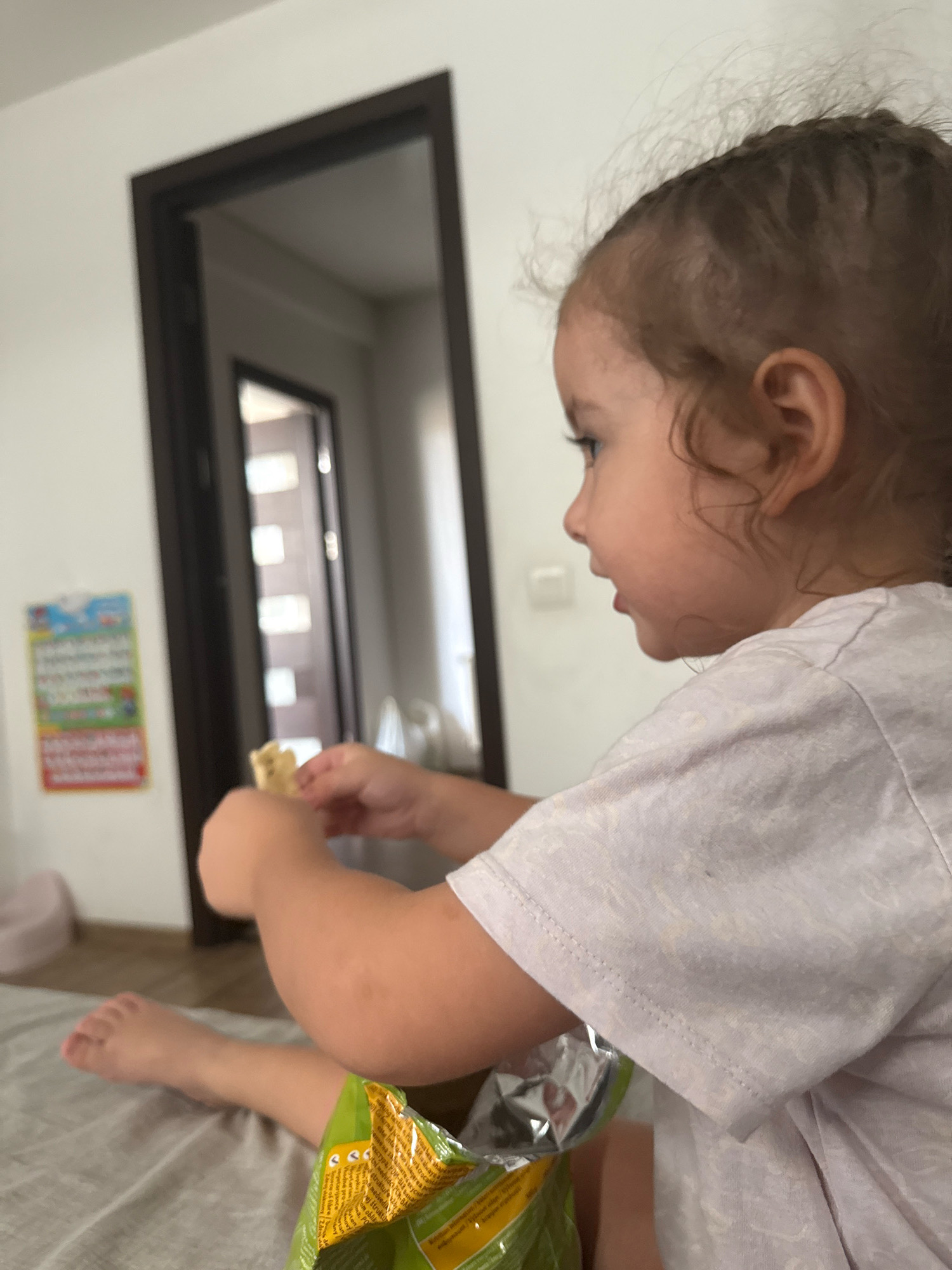 A close-up, profile shot shows a young child with curly brown hair and light skin sitting on a bed. The child is wearing a light purple t-shirt with a subtle pattern. Their right arm is bent, and their hands are holding a light-colored snack. To the left of the child, a bag of snacks is visible, partially obscuring their legs. A light gray blanket covers the bed. In the background, a dark wooden doorframe is visible, with a reflection of a hallway visible through the open doorway. A colorful children's chart is hanging on the white wall to the left of the door. The overall environment appears to be an indoor residential setting, likely a bedroom. No specific location in Chișinău, Moldova can be determined from the image.