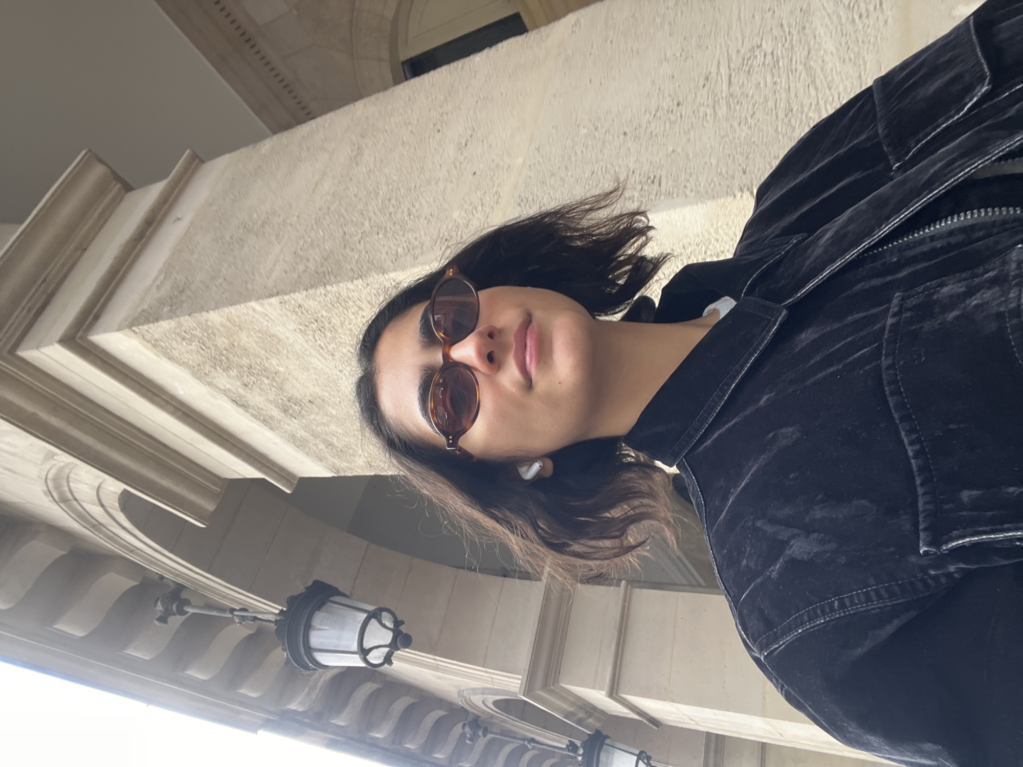 A low-angle shot captures a young woman looking upward, possibly taking a selfie, against the backdrop of an ornate stone building. She has dark, wavy, medium-length hair and is wearing tortoiseshell-framed sunglasses, with a white earbud visible in her right ear. Her expression is calm and serene, with a slight smile. She is dressed in a dark, possibly velvet or corduroy, zip-up jacket with a structured collar.
The background features the detailed, light-colored stone architecture of a classical building, consistent with structures found in Paris, France. Prominent architectural elements include decorative cornices, moldings, and an arched opening. Several traditional, lantern-style light fixtures hang from a structured ceiling, suggesting the woman is standing under an arcade, portico, or a covered walkway of a historic building.
The scene is bathed in bright natural daylight, indicating clear, sunny weather. There is no visible text in the image.
