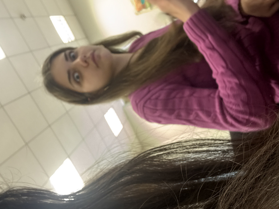The scene is an indoor environment in the city of Albi, France, characterized by an acoustic tile ceiling fitted with rectangular fluorescent light fixtures. Two individuals are present. On the far left, a portion of an individual's head and long, dark brown hair are visible, suggesting the perspective of the photographer or a very close subject. To the right, a female subject with long, light brown hair, wearing a magenta cable-knit sweater, is partially depicted. Her face is directed towards the camera, featuring wide eyes and a slightly pursed mouth, indicative of a deliberate expression. Her left arm is partially visible on the right edge of the frame. The background behind the female subject is out of focus, appearing as a light-colored surface with indistinct shapes. The lighting is artificial, originating from the overhead fixtures.