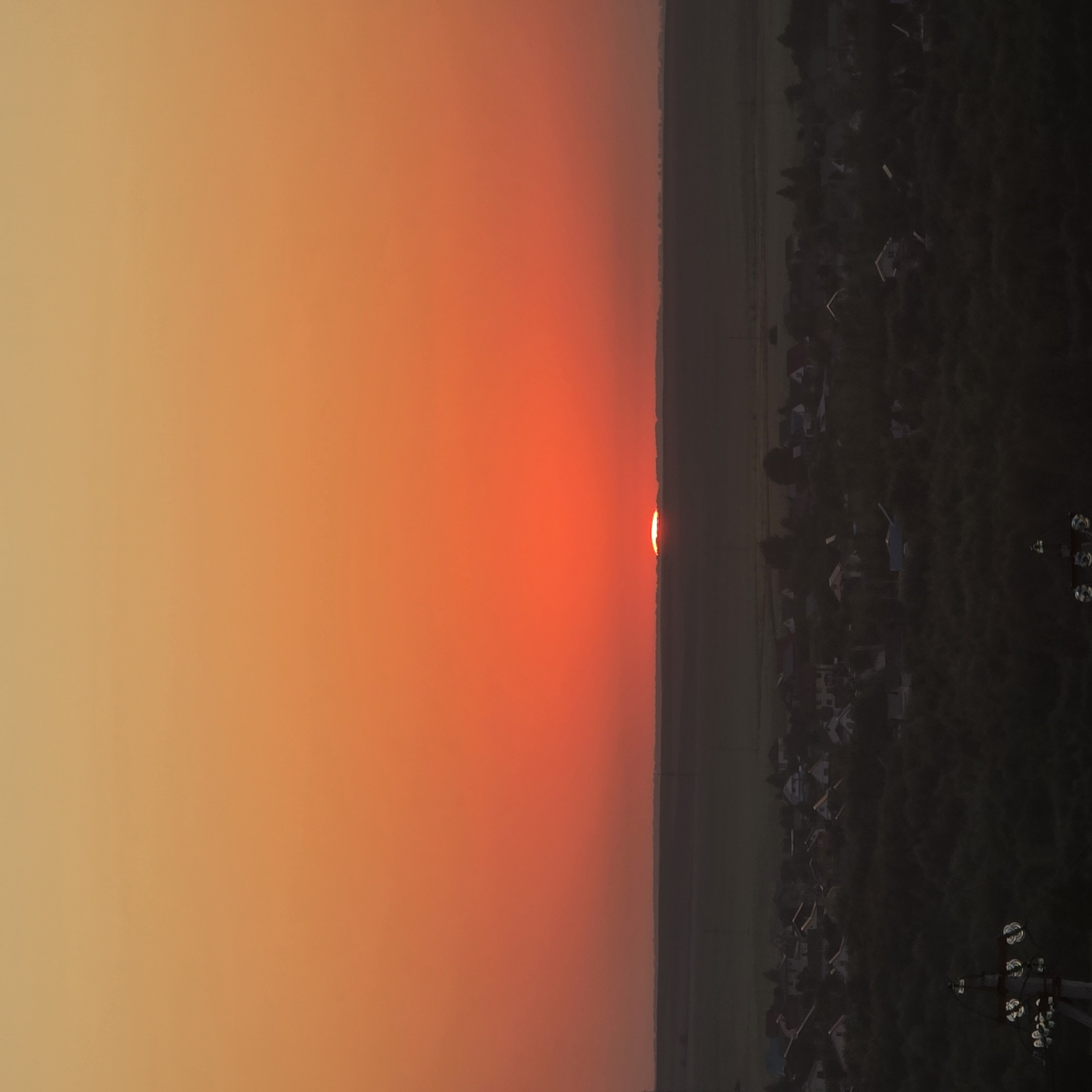 A wide-angle shot captures a sunset over a rural landscape with a town in the foreground. The sky is a gradient of orange and red, with the sun a bright sliver on the horizon. Below the sky, a dark band of land stretches across the frame, with a town nestled within it, its houses appearing as small, dark shapes. Trees and vegetation surround the town, creating a dense, dark foreground. The scene is serene and peaceful, with no signs of human activity or any identifiable landmarks. The image is devoid of any specific details that would allow for identification of the city or country.