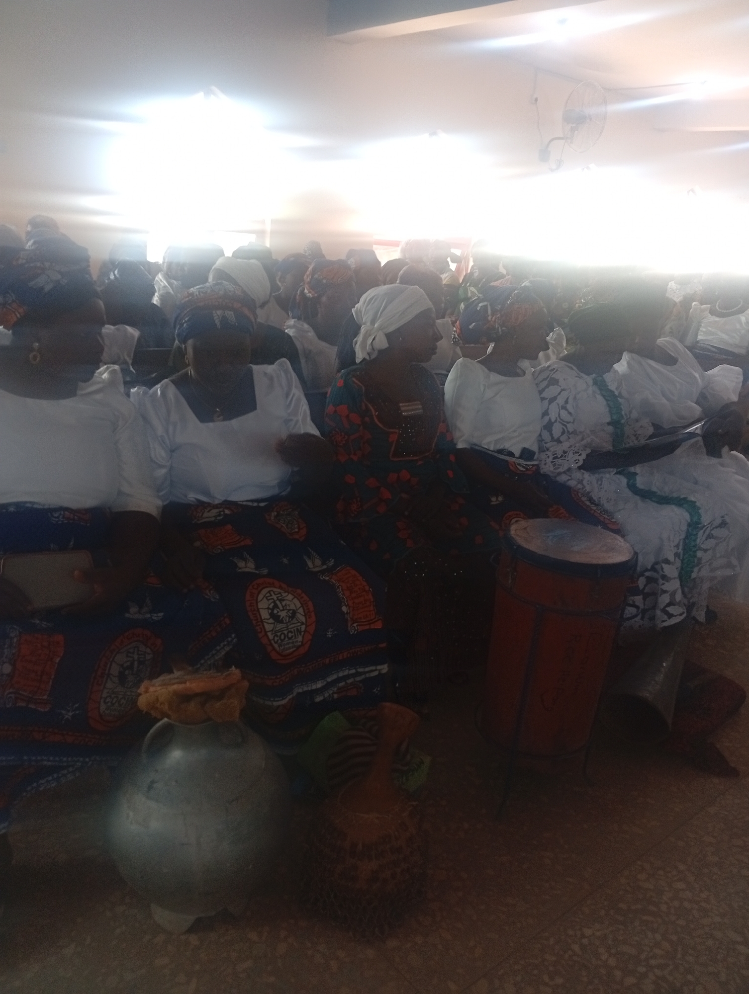 The image shows a gathering of people, predominantly women, seated in what appears to be an indoor setting. Many of the women are wearing traditional clothing, including head coverings. There is a drum and a metal pot present. The lighting is bright, possibly due to natural light coming through a window. The location is in the city of Unknown, Unknown.