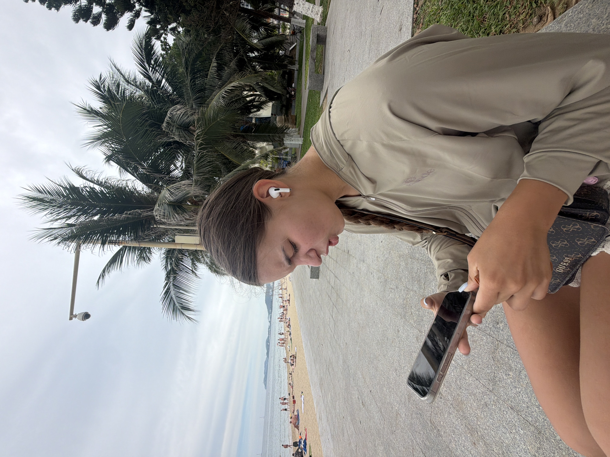 A young woman with brown hair in a braid sits on a stone bench on a paved walkway, looking at her phone. She is wearing a light olive-green hooded jacket and dark shorts. She has white wireless earbuds in her ears. A small patterned wallet with the "GUESS" logo is visible in her lap. Behind her, a sandy beach is populated with sunbathers. Beyond the beach, the ocean stretches to a distant horizon with islands. Palm trees and other foliage line the walkway, and a security camera is mounted on a pole overhead. The scene appears to be located in Nha Trang, Vietnam, a coastal city known for its beaches.