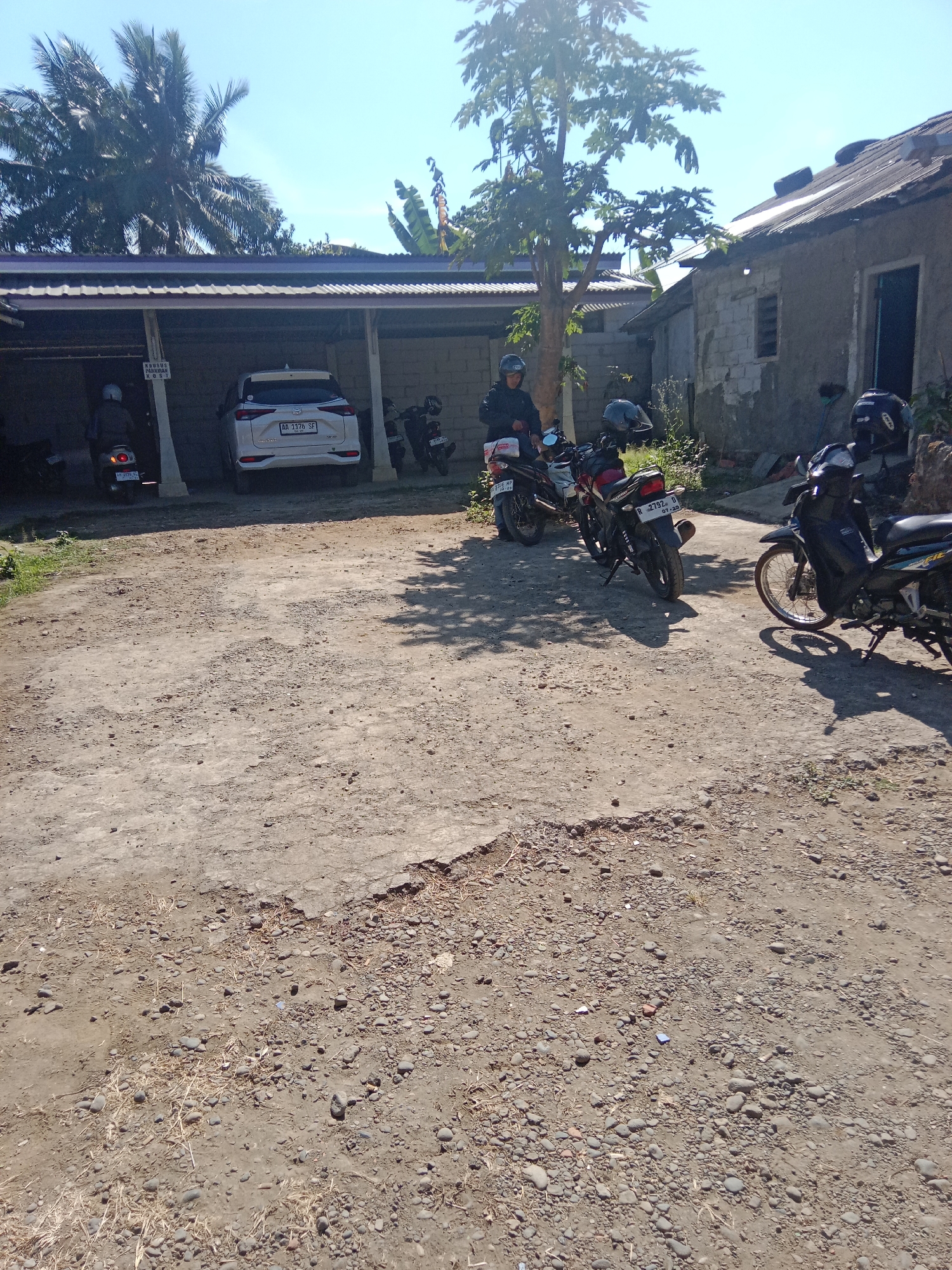 In Leksono, Indonesia, an outdoor scene is captured under a clear blue sky. The foreground is dominated by a rough, dusty ground surface with scattered stones and dried vegetation. Behind this, a paved area leads to a covered structure, likely a carport, where a white SUV with license plate "AA 3376 SF" is parked. To the left of the SUV, a person on a scooter, wearing a helmet, is partially visible. To the right of the SUV, under the carport, are several motorcycles. Outside the carport, more motorcycles are parked on the paved area. One black motorcycle in the mid-right of the image has its kickstand down. Further right, a portion of a building with a gray tiled roof and weathered walls is visible. The overall impression is of a residential or utilitarian area with multiple vehicles present.