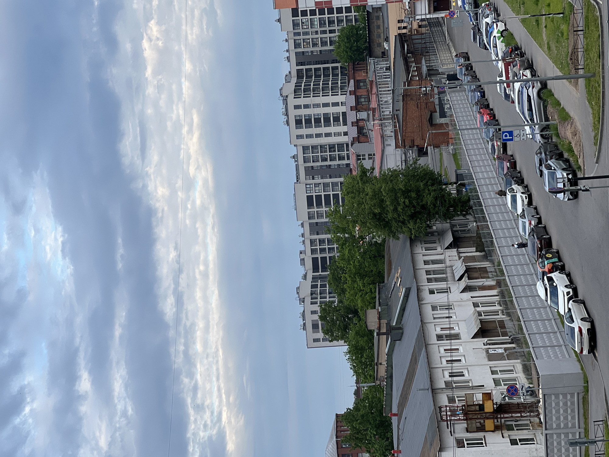 The image presents an urban landscape in Saint Petersburg, Russia, captured from an elevated perspective on a cloudy day. The scene is oriented to show a mix of sky, buildings, and a street with parked cars.
The upper left portion of the image is dominated by the sky, filled with an expanse of light blue partially obscured by large, wispy white and grey clouds. A faint, thin dark line, likely a power line, traverses the sky.
The right side of the image features various buildings and a street. In the mid-ground, a prominent row of tall, modern apartment buildings with numerous windows and balconies stands, characterized by their white and dark paneling. Interspersed among these and on the rooftops of lower structures are several green trees, adding touches of nature to the urban fabric.
Below the modern high-rises are several lower-rise buildings, some displaying older architectural styles with brick facades, while others are painted white. A complex arrangement of fences, staircases, and industrial-like structures can be seen between and around these buildings.
In the foreground, a paved road winds diagonally across the lower right. It is densely lined with numerous parked passenger cars of various models and colors, including white, grey, and darker hues. A grassy verge and a sidewalk run alongside the road. Several road signs are visible: a prominent blue square parking sign with a white 'P' and a wheelchair symbol, accompanied by the text "5m," and another red circular "no stopping" sign (red diagonal line on a blue background). A single individual, dressed in a dark top and light trousers, is discernible on the sidewalk near the parked cars.
The lighting suggests it is daytime, possibly an overcast afternoon, with no harsh shadows visible. The overall scene depicts a typical, bustling urban street, reflecting the daily life and architectural diversity common in a large city like Saint Petersburg.