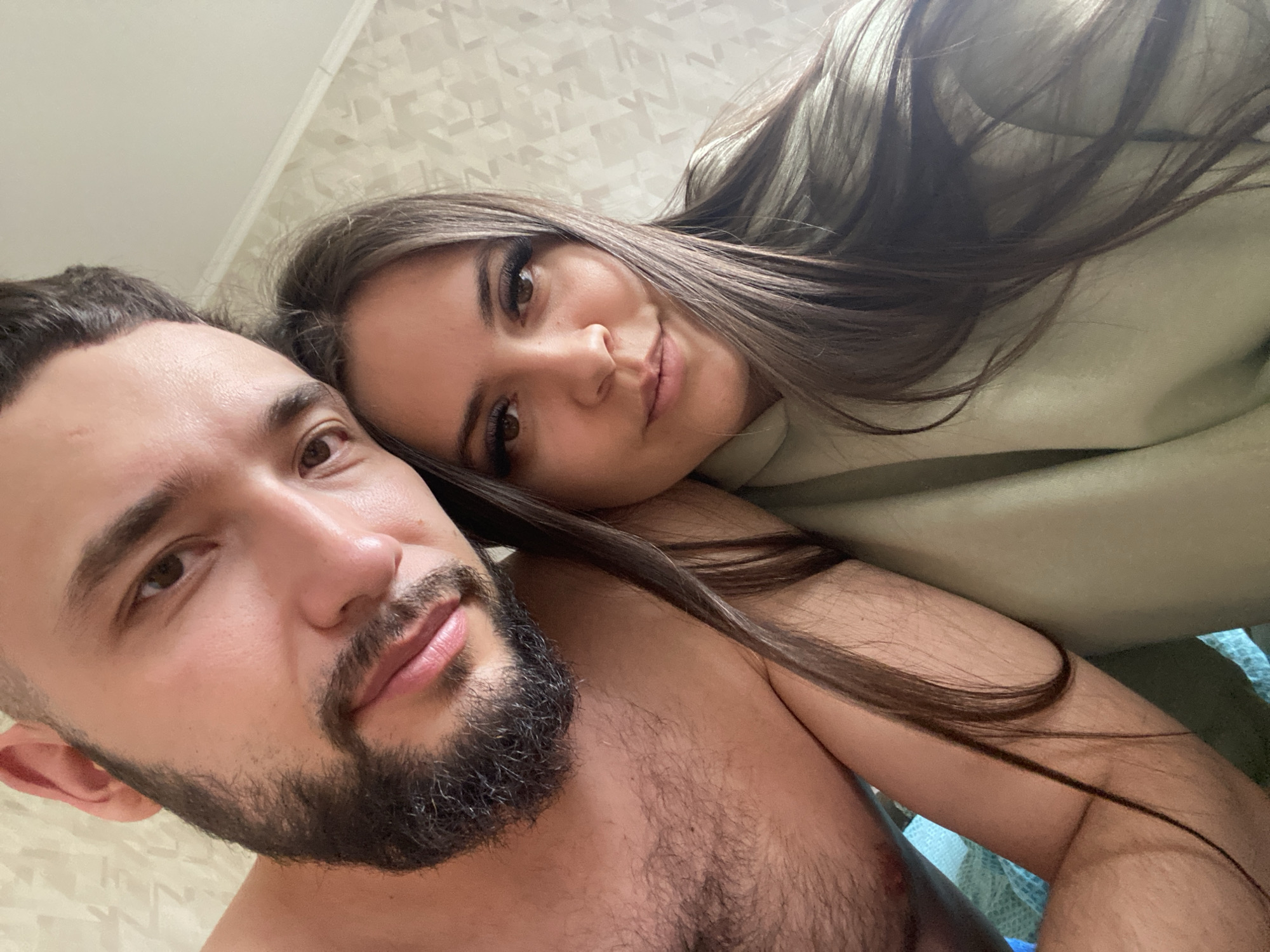 A close-up captures a man and a woman posing for a selfie. The man, in the foreground, displays a dark beard and chest hair, while the woman, situated behind him, rests her head on his shoulder. She has long, dark hair and appears to be wearing a light green sweatshirt. The backdrop features a patterned wall. No information is available from the image to suggest that this image was taken in Zhigalki, Russia.