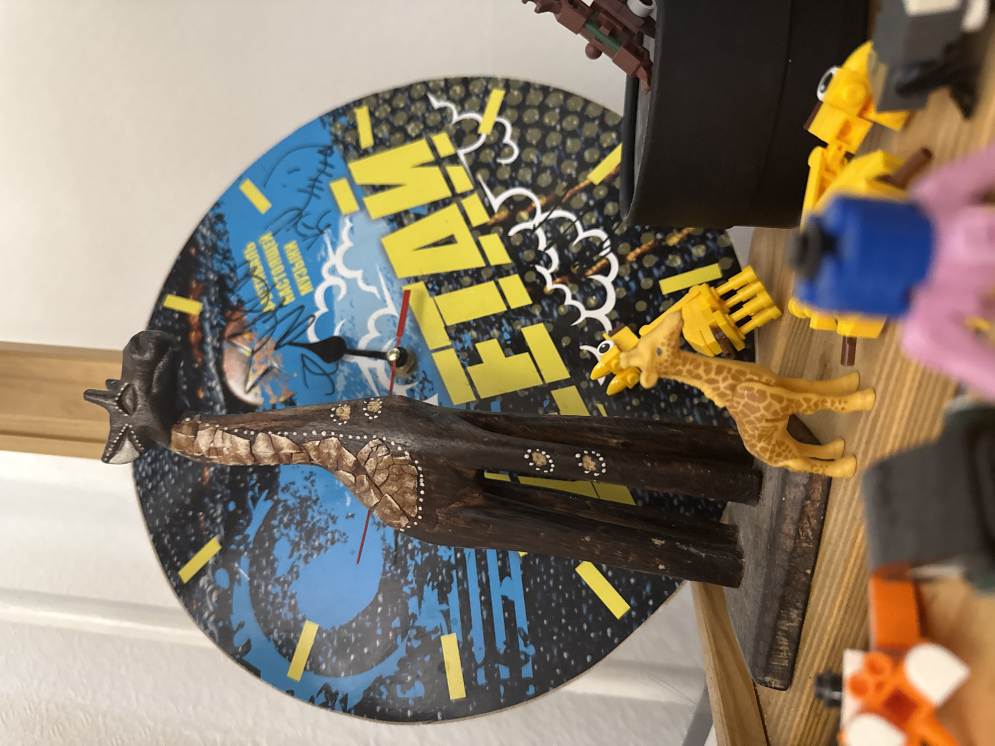 A close-up shot shows a cluttered tabletop with various objects. In the center is a colorful, round wall clock with a blue and black design, with yellow lines denoting the hours. The word "FLAM!" is prominently displayed in large yellow letters. A dark brown wooden giraffe statue stands in front of the clock. A plastic yellow giraffe toy is positioned next to the wooden giraffe. In the upper right, there are Lego pieces of various colors including yellow, orange, pink, blue, and black. The table appears to be made of light-colored wood. There is no visual evidence to confirm the location as Chaikovsky, Russia.