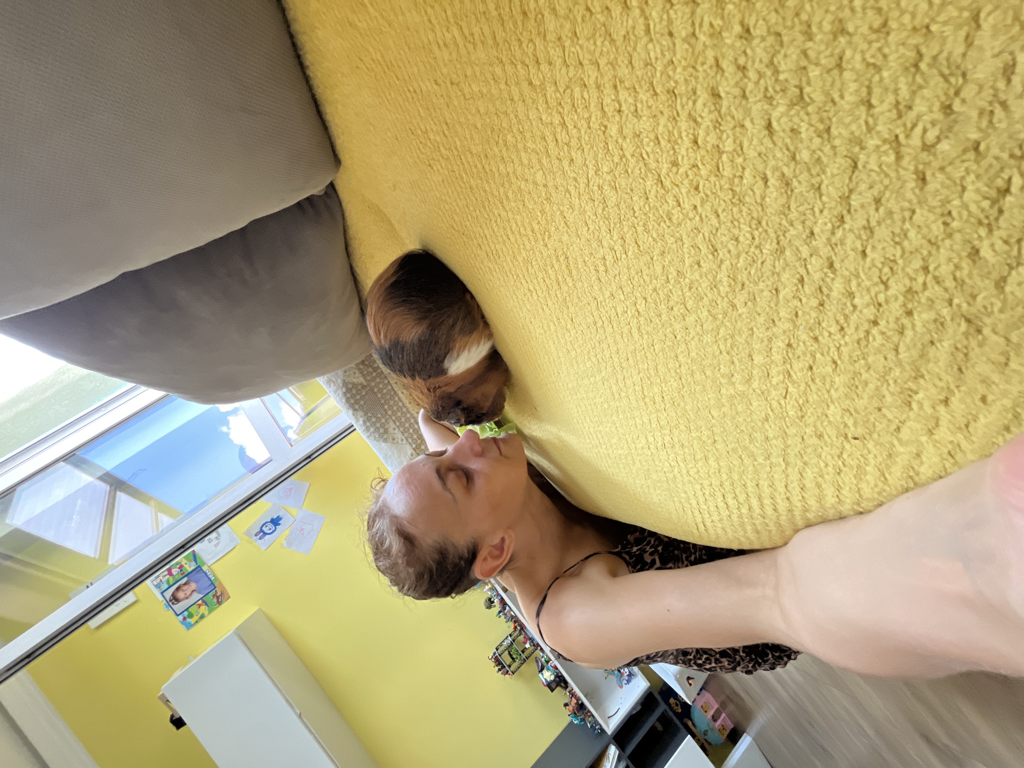 An overhead shot shows a woman with her eyes closed lying on a yellow textured surface next to a guinea pig. The guinea pig has brown, white, and black markings and is nuzzling the woman's face. The woman has her arms wrapped around herself and is wearing a leopard print top with thin straps. In the background, a yellow wall is visible, adorned with various pictures and drawings. A window with a view of the sky and clouds is also present, along with a shelf unit filled with toys and books. The scene appears to be indoors, in a home environment.