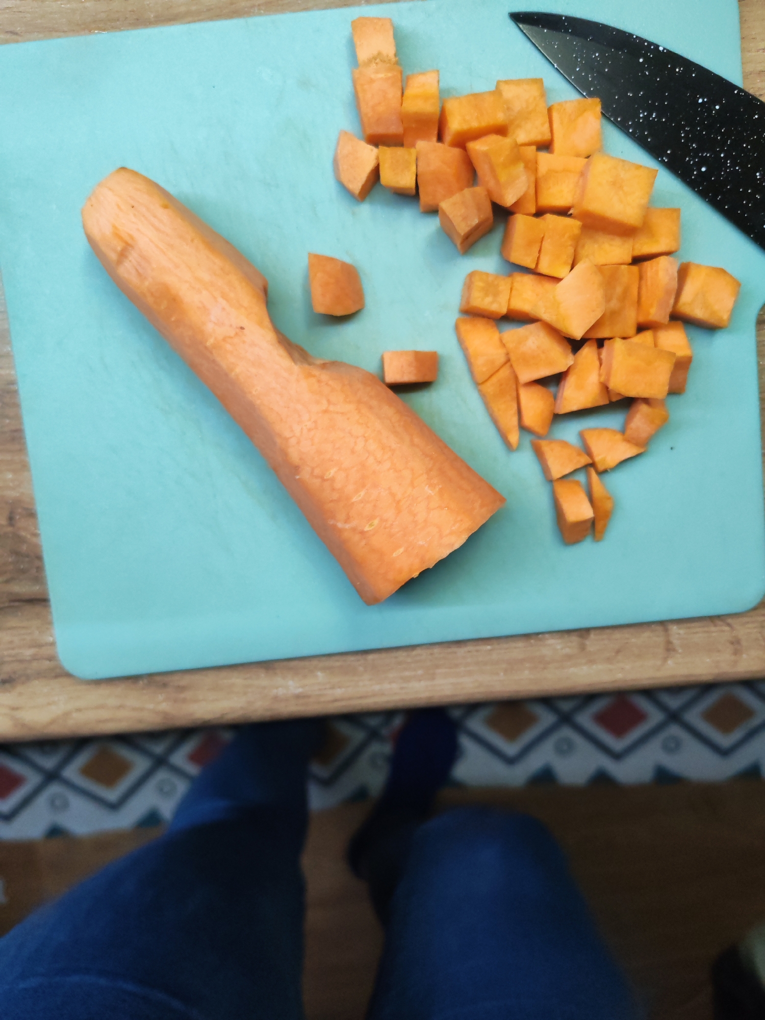 A still life shows an orange carrot, partially diced, on a light blue cutting board. A black handled knife with a spotted blade sits nearby. The cutting board rests on a wood-grained surface with a patterned rug or floor visible underneath. A person wearing dark blue jeans and socks is standing, presumably holding the camera. The image was taken indoors. There is no evidence to suggest that this image was taken in Izhevsk, Russia.