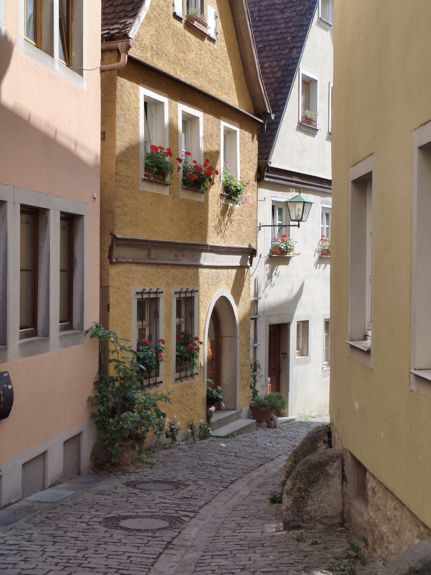 The image shows a narrow cobblestone street in Rothenburg ob der Tauber, Germany, lined with historic buildings. The buildings have a mix of yellow and cream facades, with steeply pitched roofs covered in reddish-brown tiles. Flower boxes with red geraniums are visible on window sills, adding pops of color. On the left, a building with a peach-colored facade features tall, narrow windows. The street is paved with rough cobblestones, and two circular manhole covers are visible. A large, moss-covered boulder sits on the right side of the street, next to a stone foundation. The scene is illuminated by sunlight, casting shadows across the street and buildings. No people are present in the image.