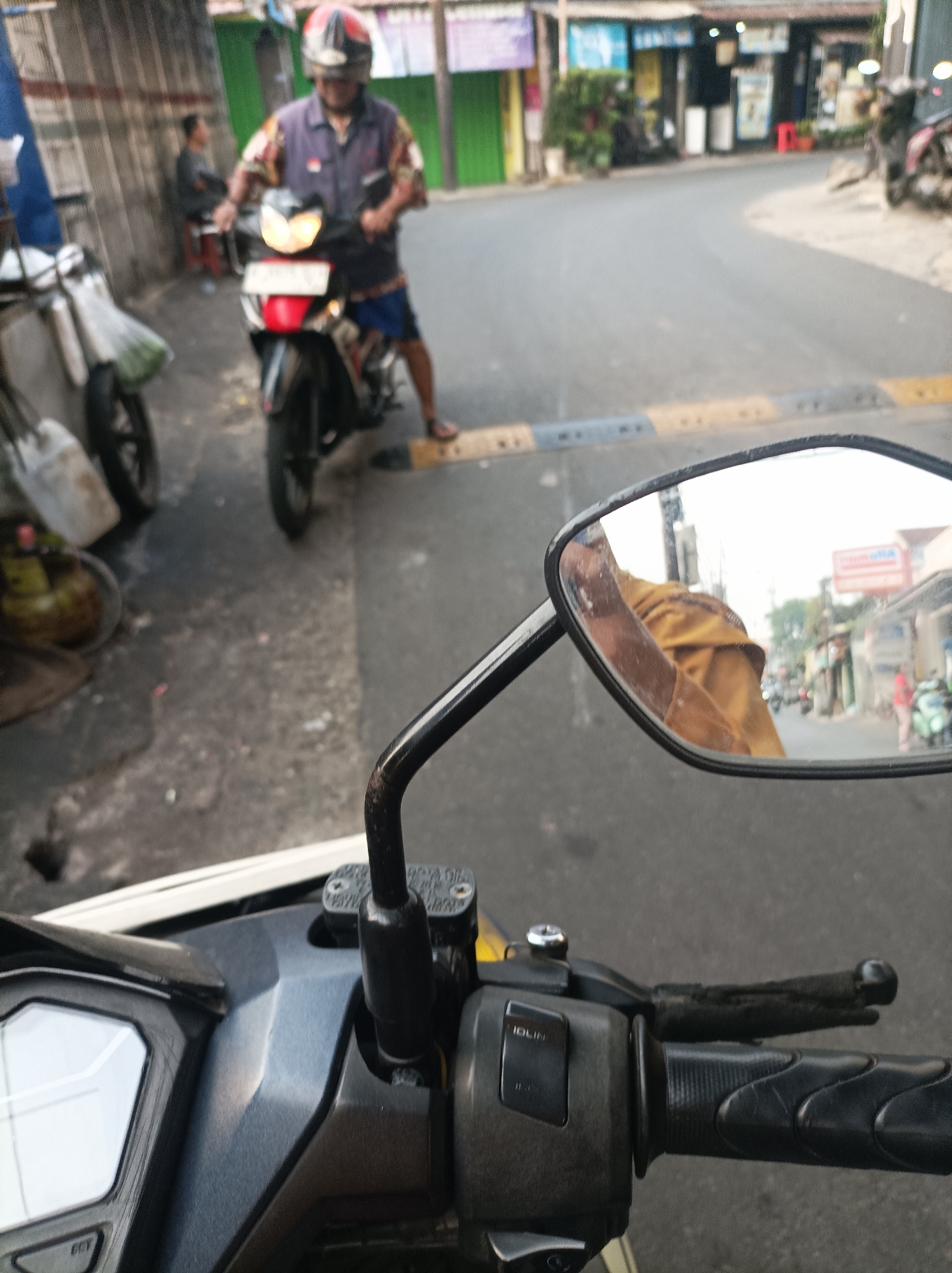 The image captures a street scene in West Jakarta, Indonesia, from the perspective of someone riding a motorcycle. In the foreground, a motorcycle handlebar and dashboard are visible. The rider's hand, wearing a brown garment, is reflected in the motorcycle's rearview mirror. In the middle ground, a man wearing a helmet and a colorful shirt is riding another motorcycle. He is in motion, heading away from the camera. To the left, a person is seated on a stool near a food cart. The background shows a street lined with buildings and other vehicles, including motorcycles. The overall impression is of a typical urban environment in Jakarta during daytime.