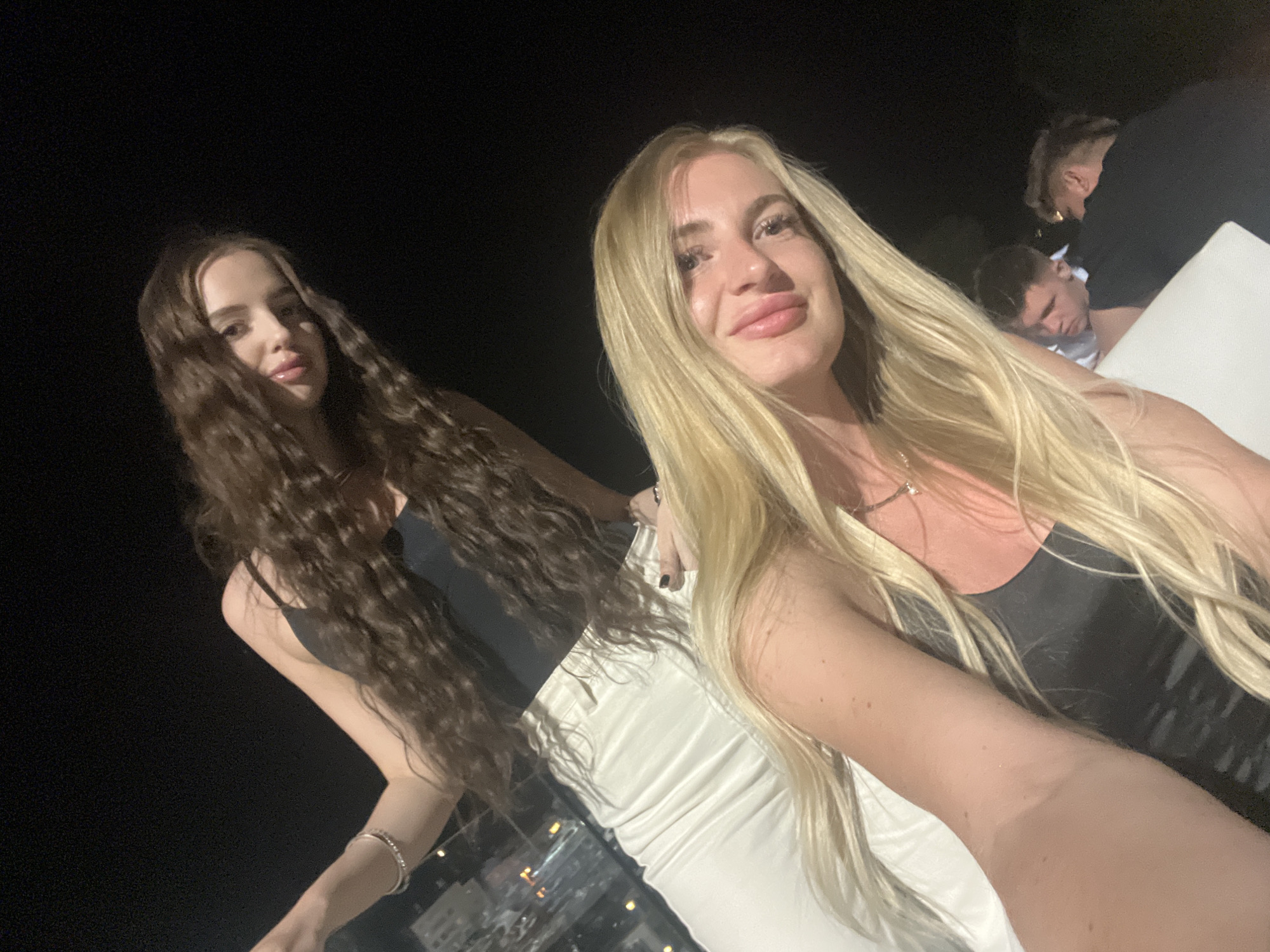 The image shows two young women posing together at night in what appears to be a high-altitude location, possibly a rooftop bar or balcony, overlooking a city. The city in the background is likely Budva, Montenegro, based on the urban layout and coastal environment.
The woman on the right has long, blonde hair and is wearing a dark top. She is holding the camera for a selfie. The woman on the left has long, curly brown hair and is wearing a dark top and white pants. Both women are fair-skinned and have makeup.
In the background, there are two men, one appearing to be asleep or resting on a white surface, the other standing nearby. The overall scene suggests a nighttime social gathering. The environment is dimly lit, with city lights visible in the distance.