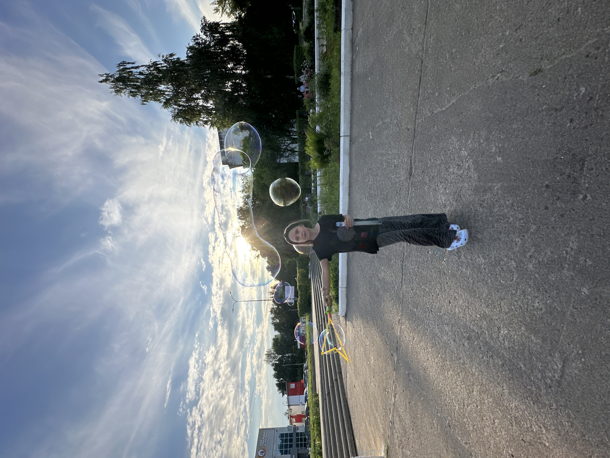 A young person is standing on an asphalt surface, holding a bubble wand with a large set of bubbles floating in front of them. The person is wearing a dark t-shirt and patterned pants, with white shoes. Behind them, there are stairs leading up to a building with a sign that says "МИР ОДЕЖДЫ". Trees and a lamppost are visible in the background, as well as other people sitting on a bench in a park-like setting. The sky is blue with wispy clouds, and the sun is shining, creating lens flare. The scene appears to be in a public outdoor space, likely a park or square, in the city of Chaikovsky, Russia.