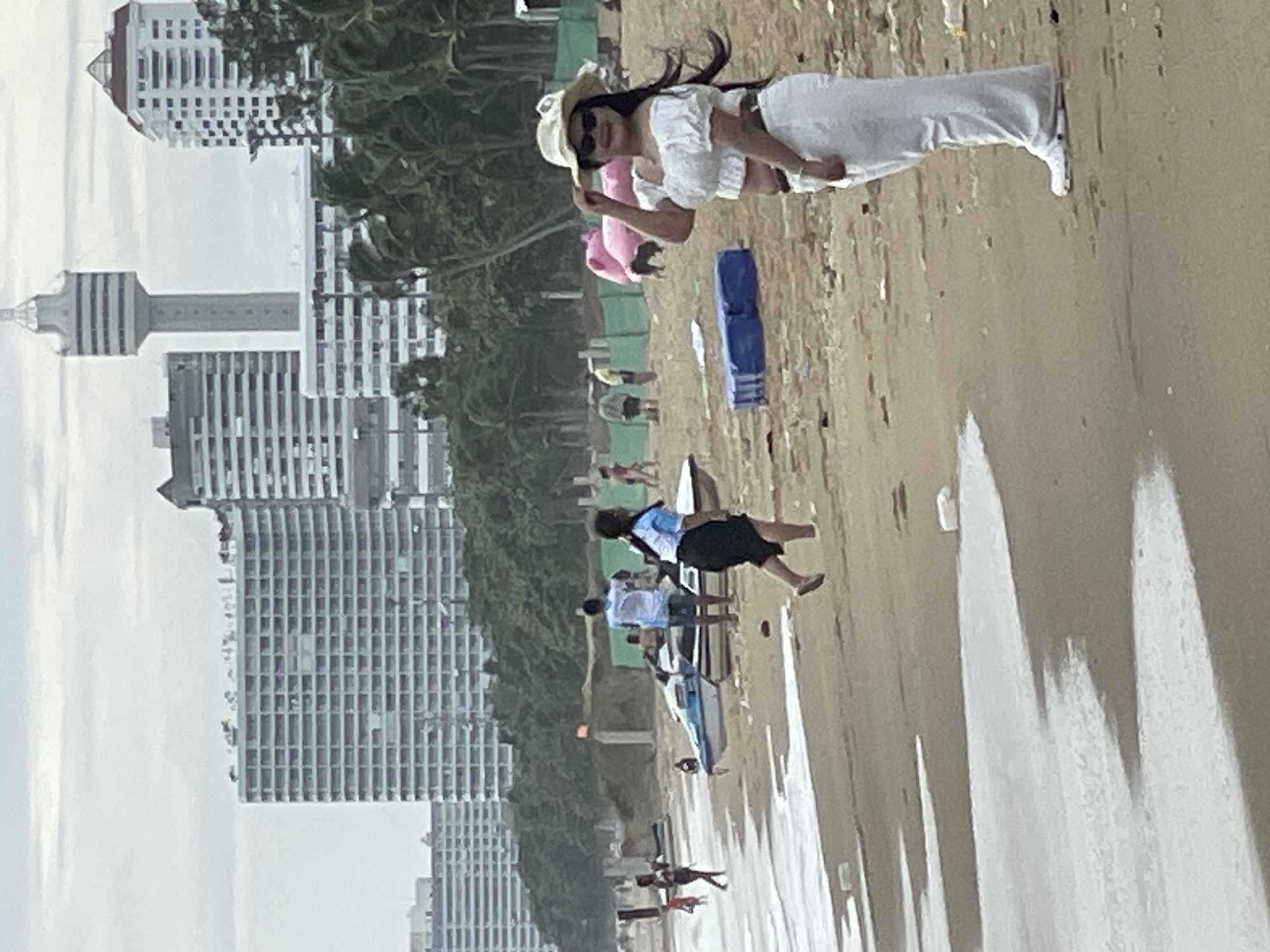 A group of people are seen on the sandy beach in Pattaya City, Thailand, with the cityscape in the background. A woman in the foreground is wearing a white off-the-shoulder top and a white skirt, and is holding a straw hat. Behind her, several other individuals are scattered on the beach, some walking and others standing. A jet ski is visible near the water's edge. In the distance, numerous high-rise buildings dominate the skyline, indicative of Pattaya's urban development. The sky is overcast.